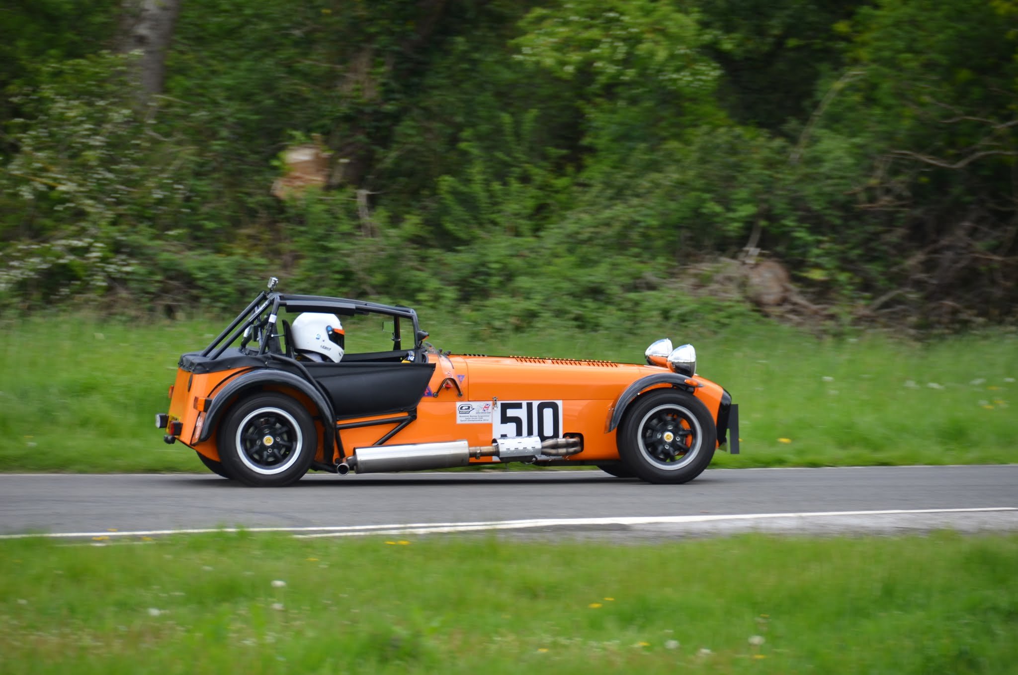 An orange Caterham race car with the number 510 drives along a paved road surrounded by green grass and trees.
