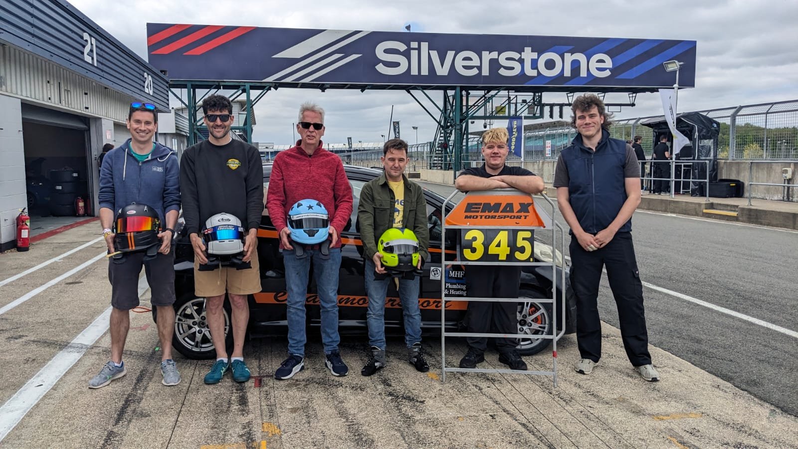 Group of seven men standing in front of a race car at a racing circuit, each holding a helmet, with a large Silverstone sign overhead.