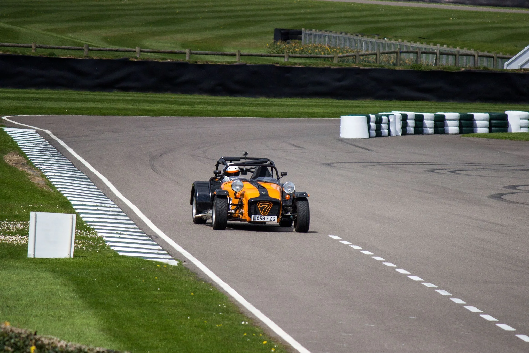 A orange and black open-wheel Caterham race car on a race track with green grass and safety barriers.