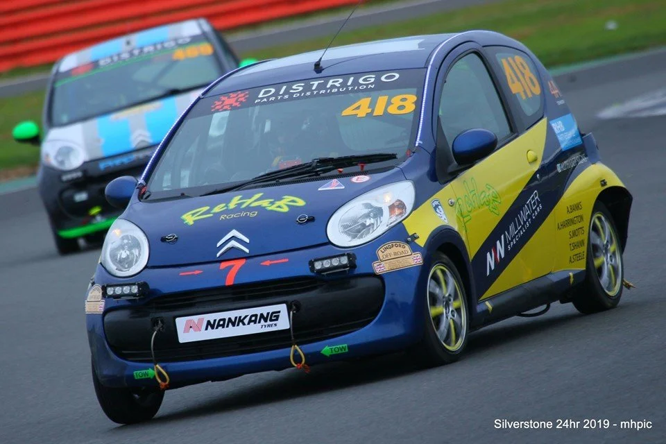A race car with a blue and yellow color scheme, number 418, on a racetrack with another car in the background.