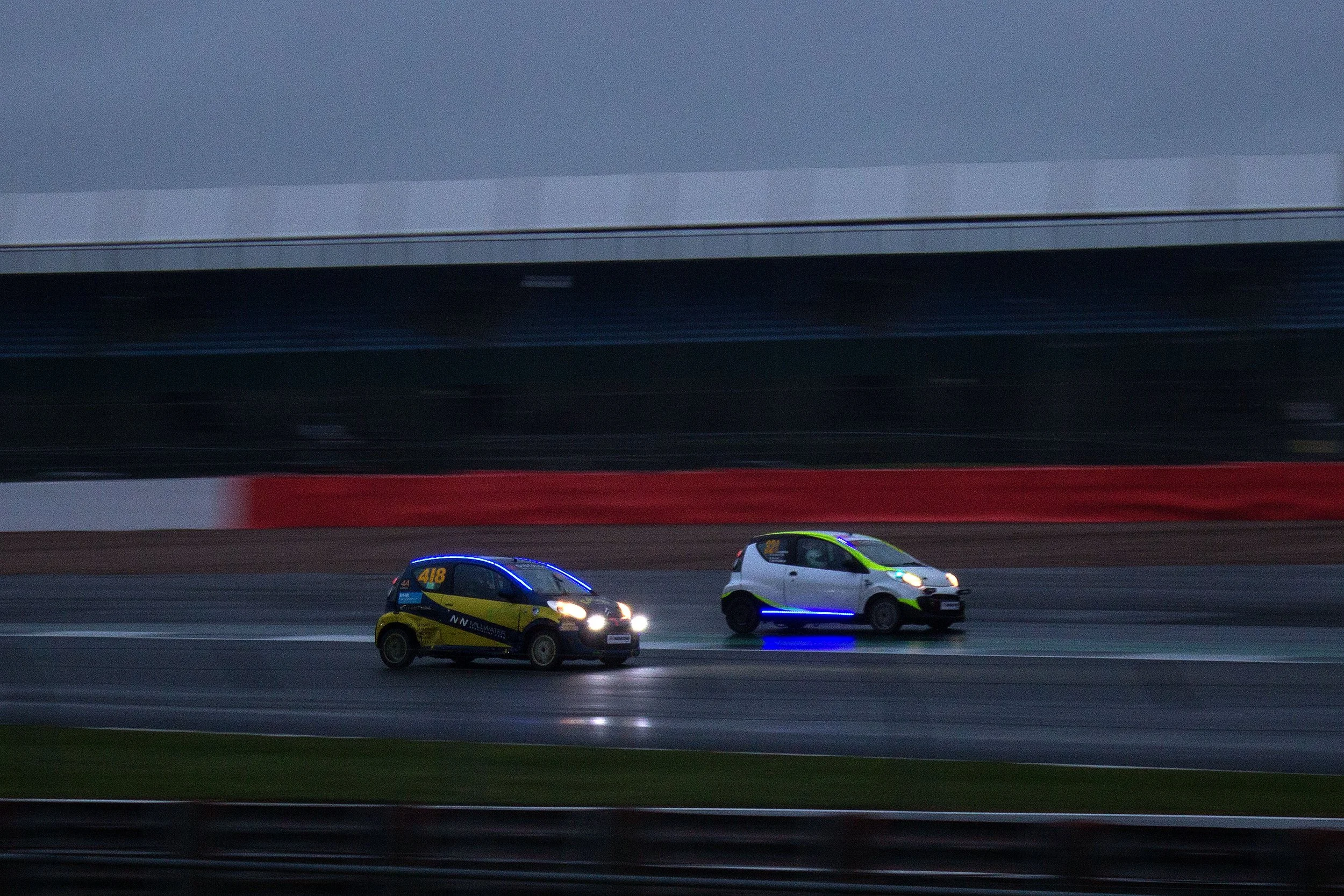 Two small race cars on a wet racetrack during dusk, with one car displaying neon blue lighting accents.