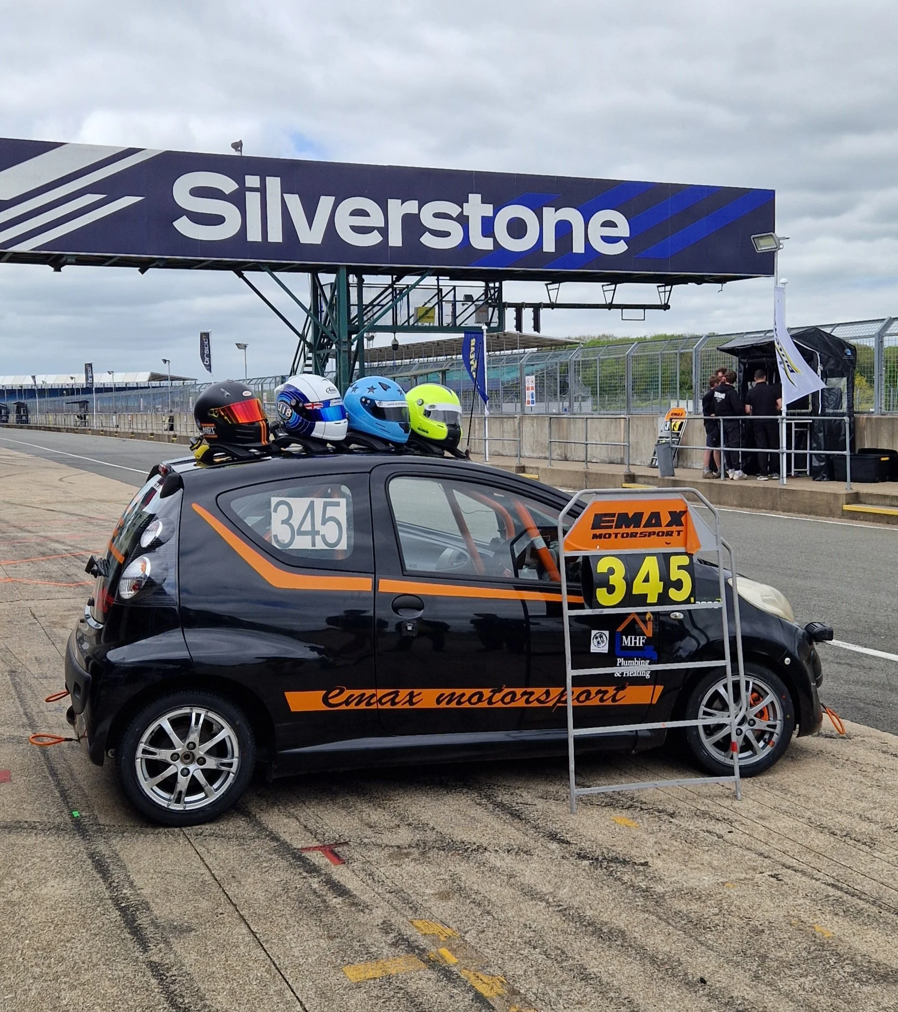 A small black race car with orange and yellow accents, labeled '345' and 'emax motorsport,' parked in a race track pit lane with four racing helmets on the roof, three helmets are blue, green, and black. In the background, there is a large sign readi