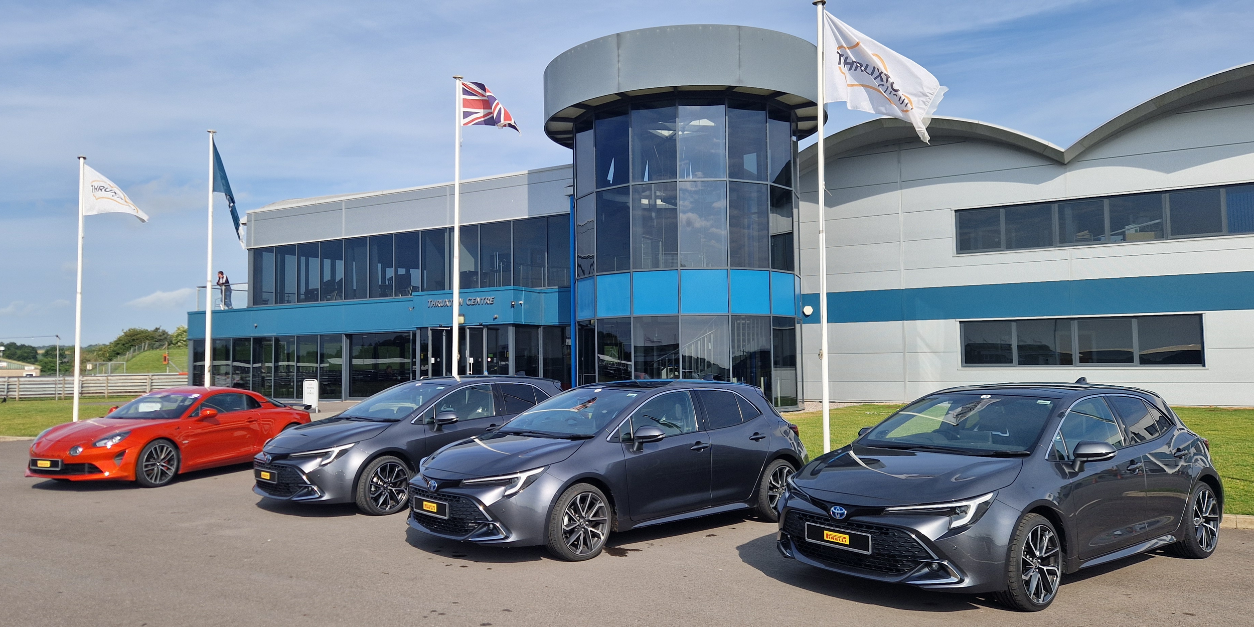 Four cars parked in front of a modern blue and gray building with flags flying on poles.