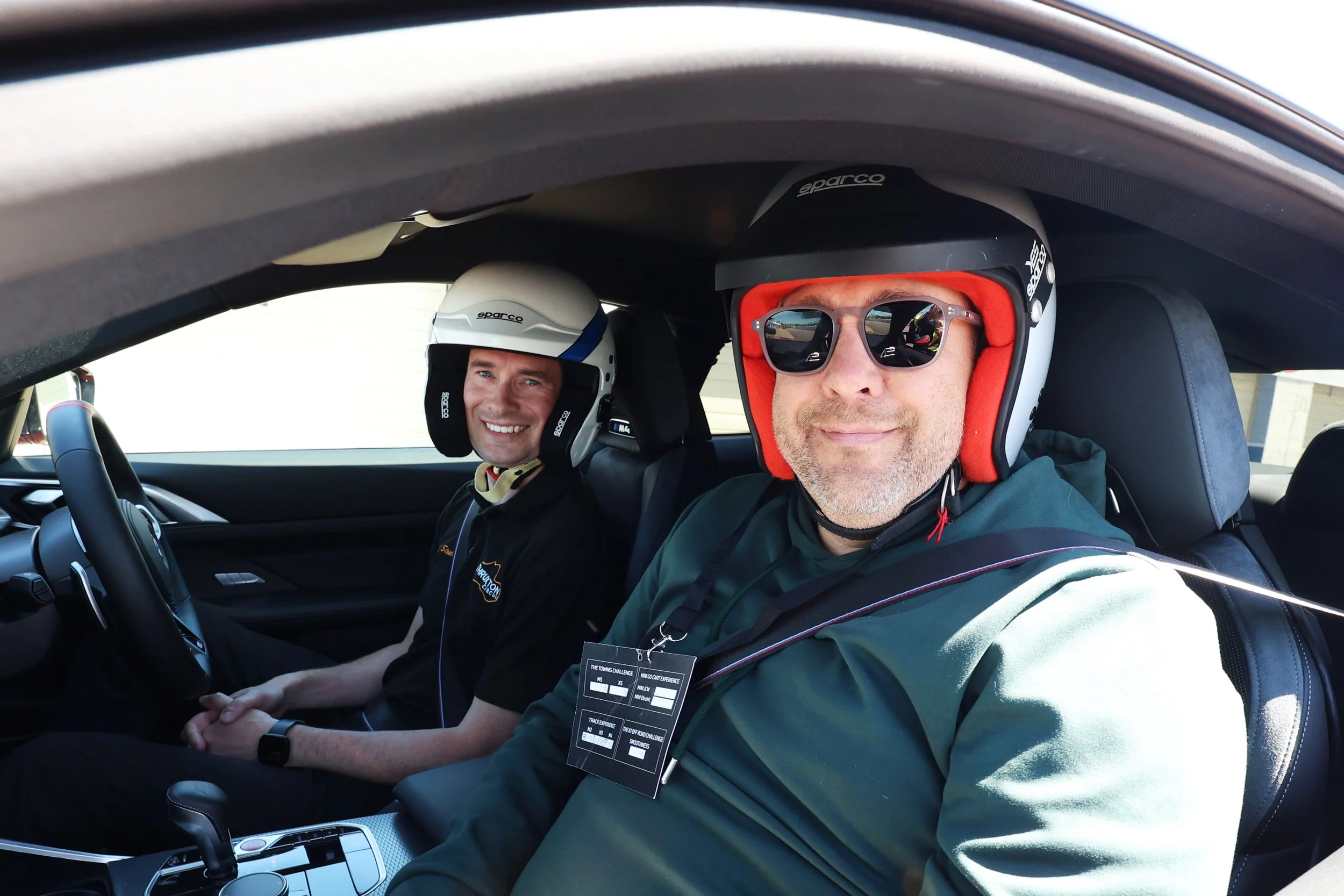Two men sit inside a car, both wearing racing helmets and sunglasses, smiling at the camera. The driver has a black helmet with a sparco logo, and the passenger has a white helmet with a sparco logo.