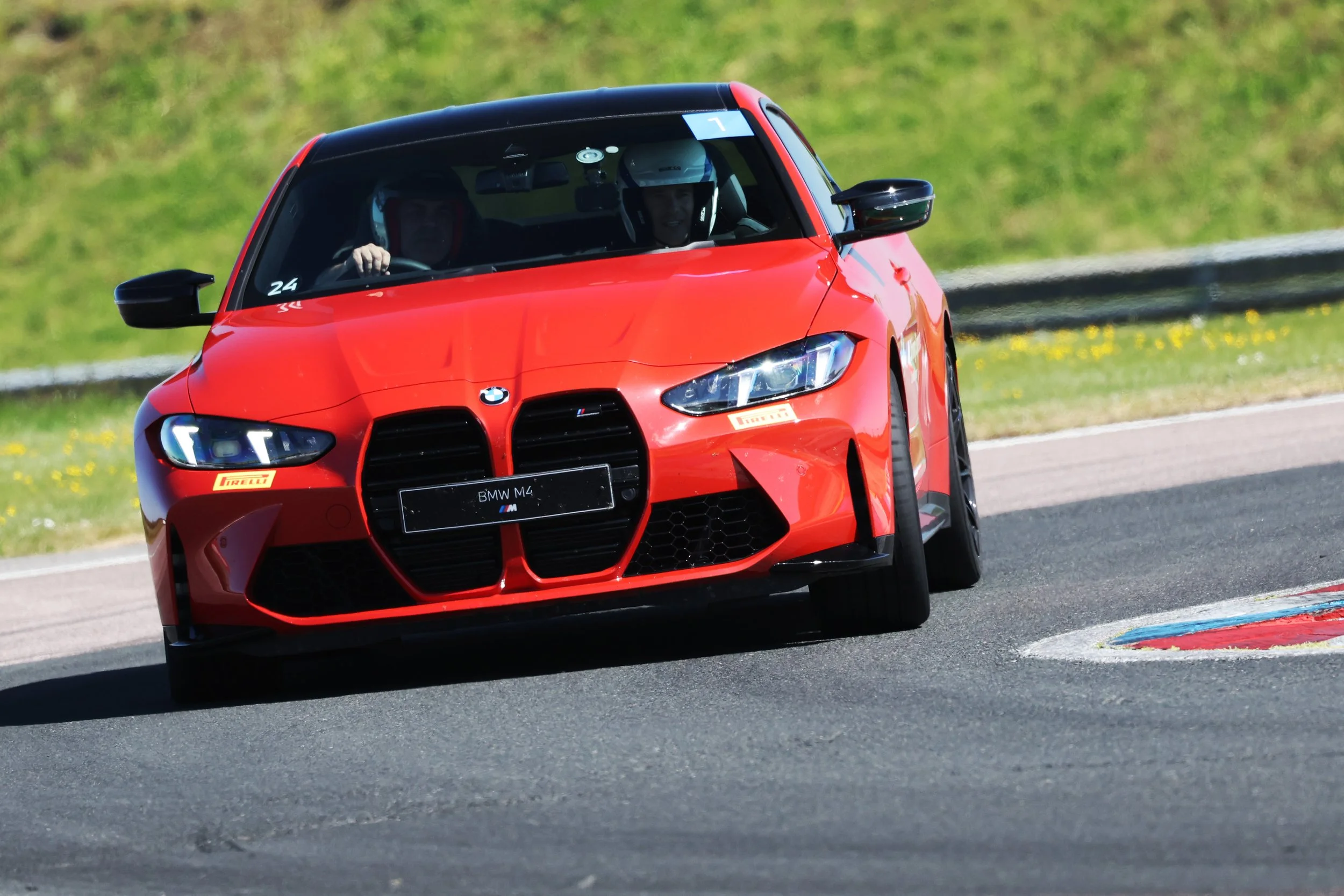 Red BMW M4 race car on a track with two drivers wearing helmets, navigating a turn on a race track with green grassy surroundings.