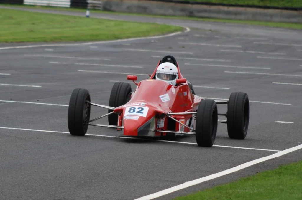 A red vintage open-wheel race car on a racetrack with the number 82 on its nose and a driver wearing a white helmet.