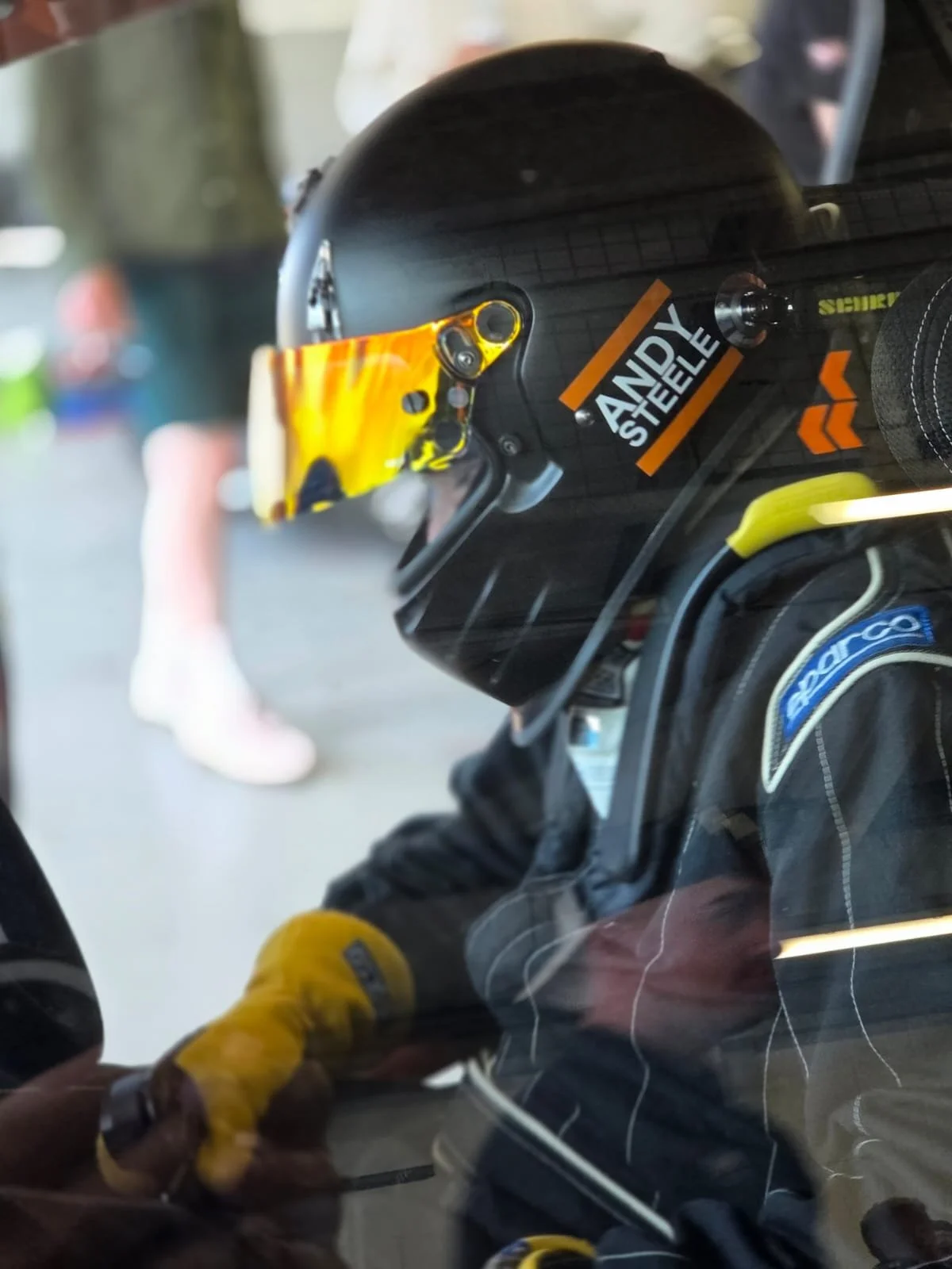 Race car driver wearing a black helmet with stickers, including one with the name 'Andy Steele', and yellow gloves, sitting in the cockpit of a race car.