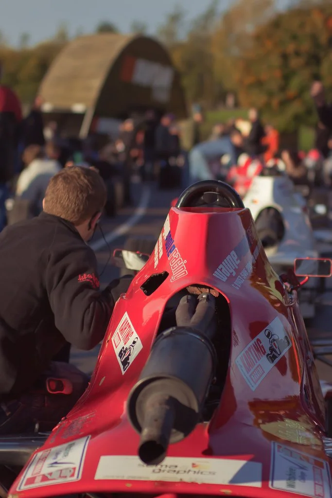 Close-up of a red race car cockpit with a large black exhaust pipe in the foreground and a person working on it, surrounded by a busy outdoor event with people and cars in the background.