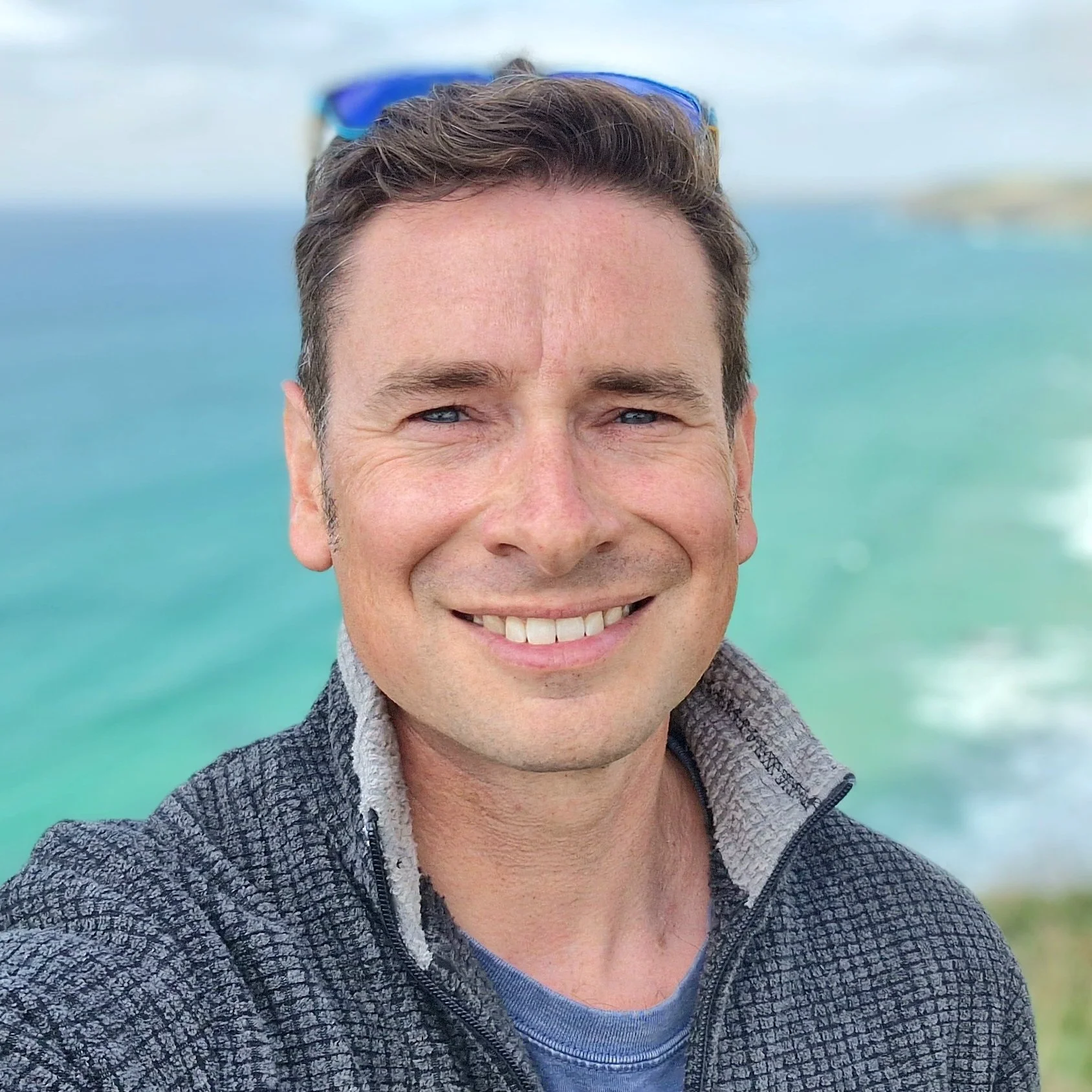 Andy Steele, smiling, with sunglasses on his head and wearing a zip-up jacket.