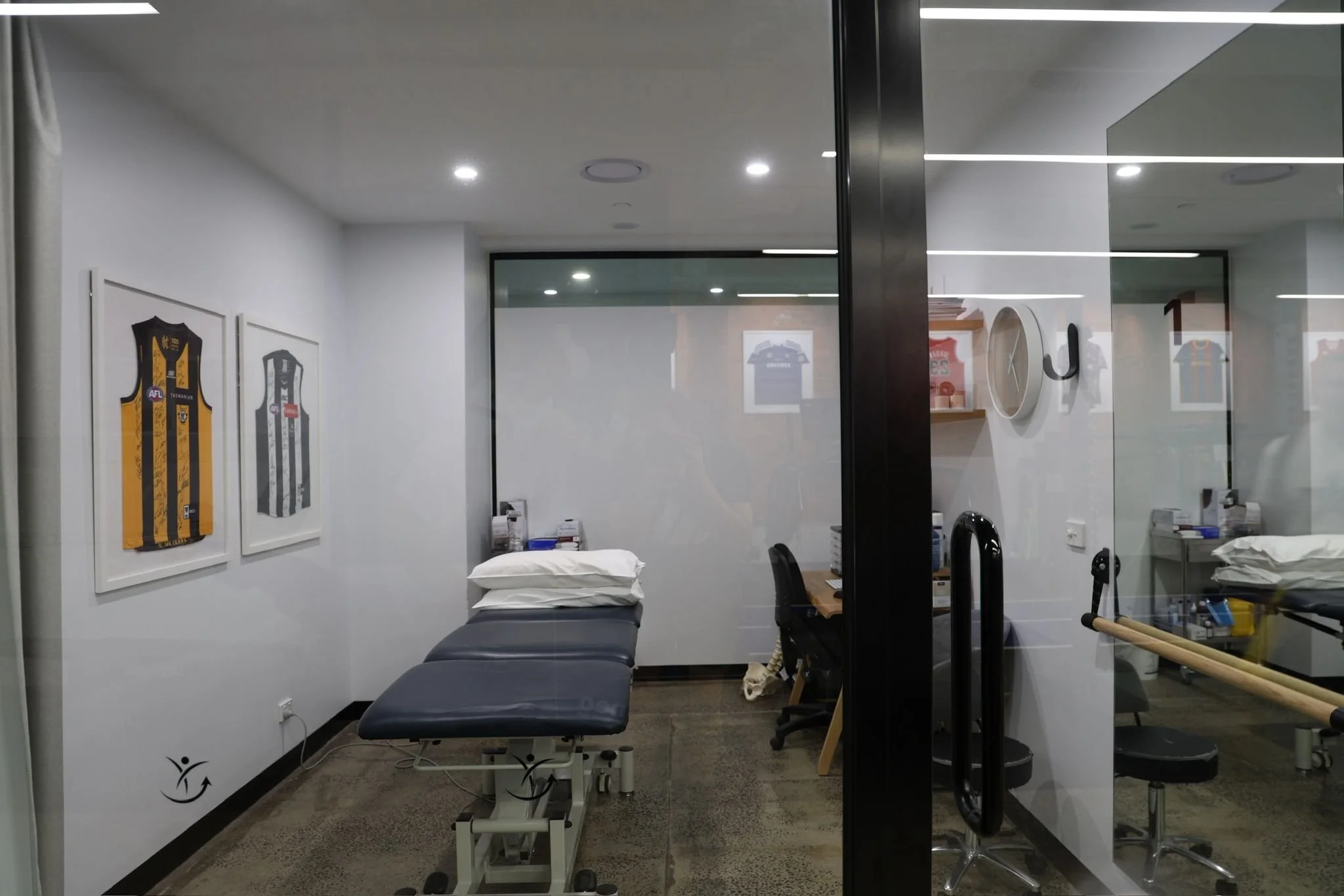 A medical examination room seen through a glass door. The room has framed sports jerseys on the wall and contains a treatment table, medical supplies, and chairs.
