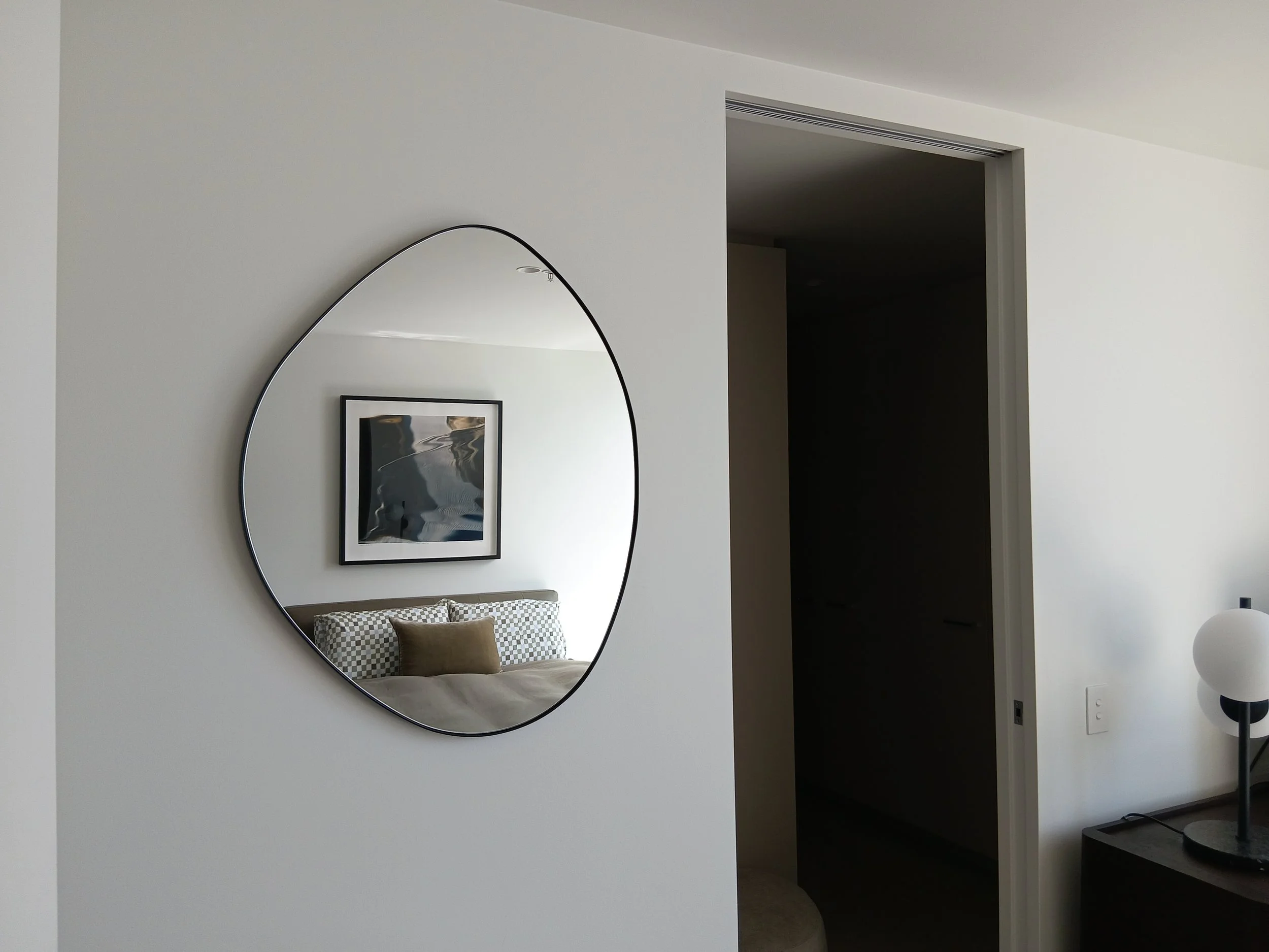 Modern bedroom with a large oval mirror on the wall, reflecting a bed with checkered bedding and a brown pillow, a framed black-and-white photo, and part of a chair. An open doorway leads to a dark room. A white lamp with a black stand is partially v