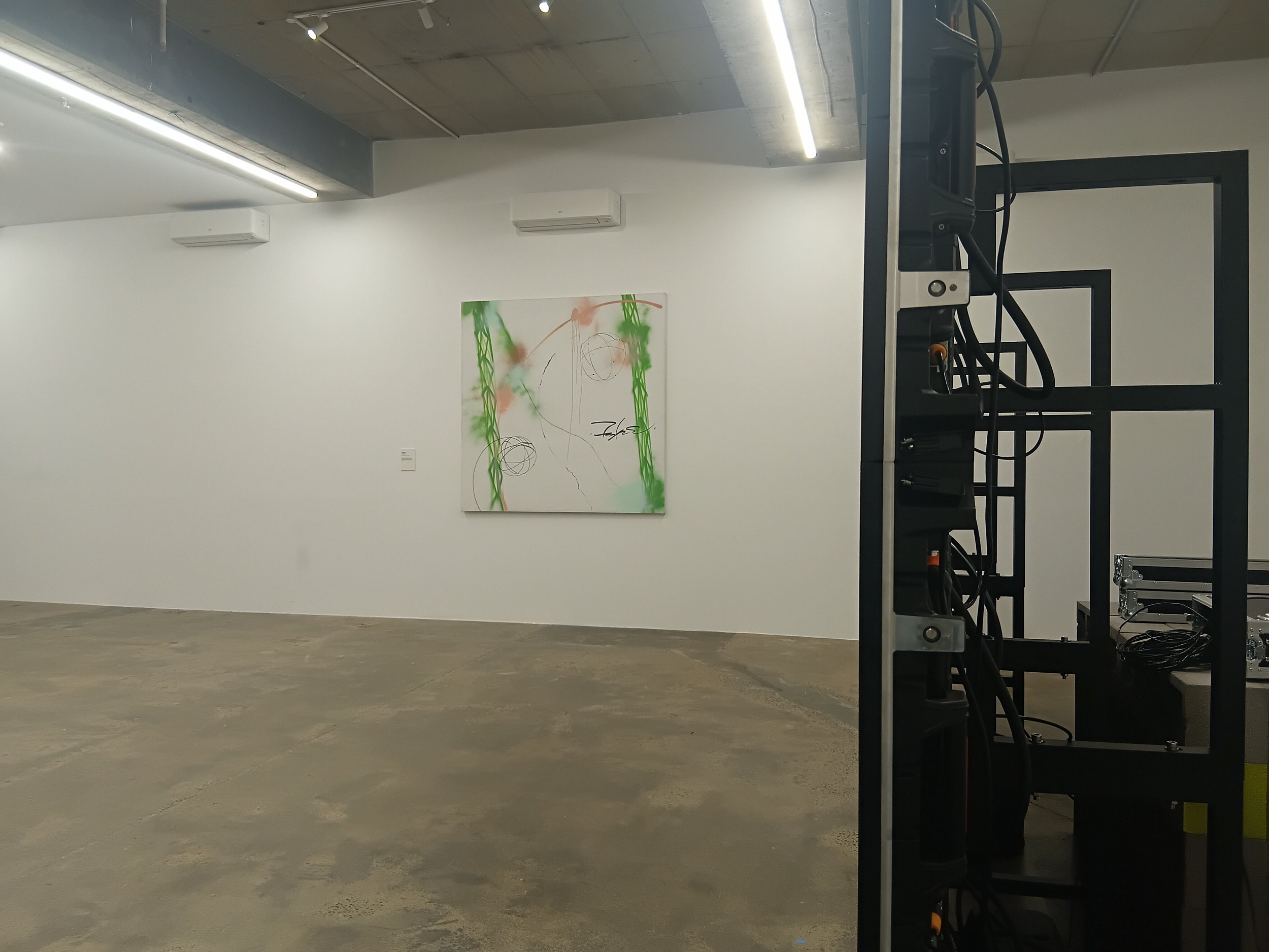 An art gallery with a white wall featuring colorful abstract artwork and a sculpture with wires and metal frames in the foreground.