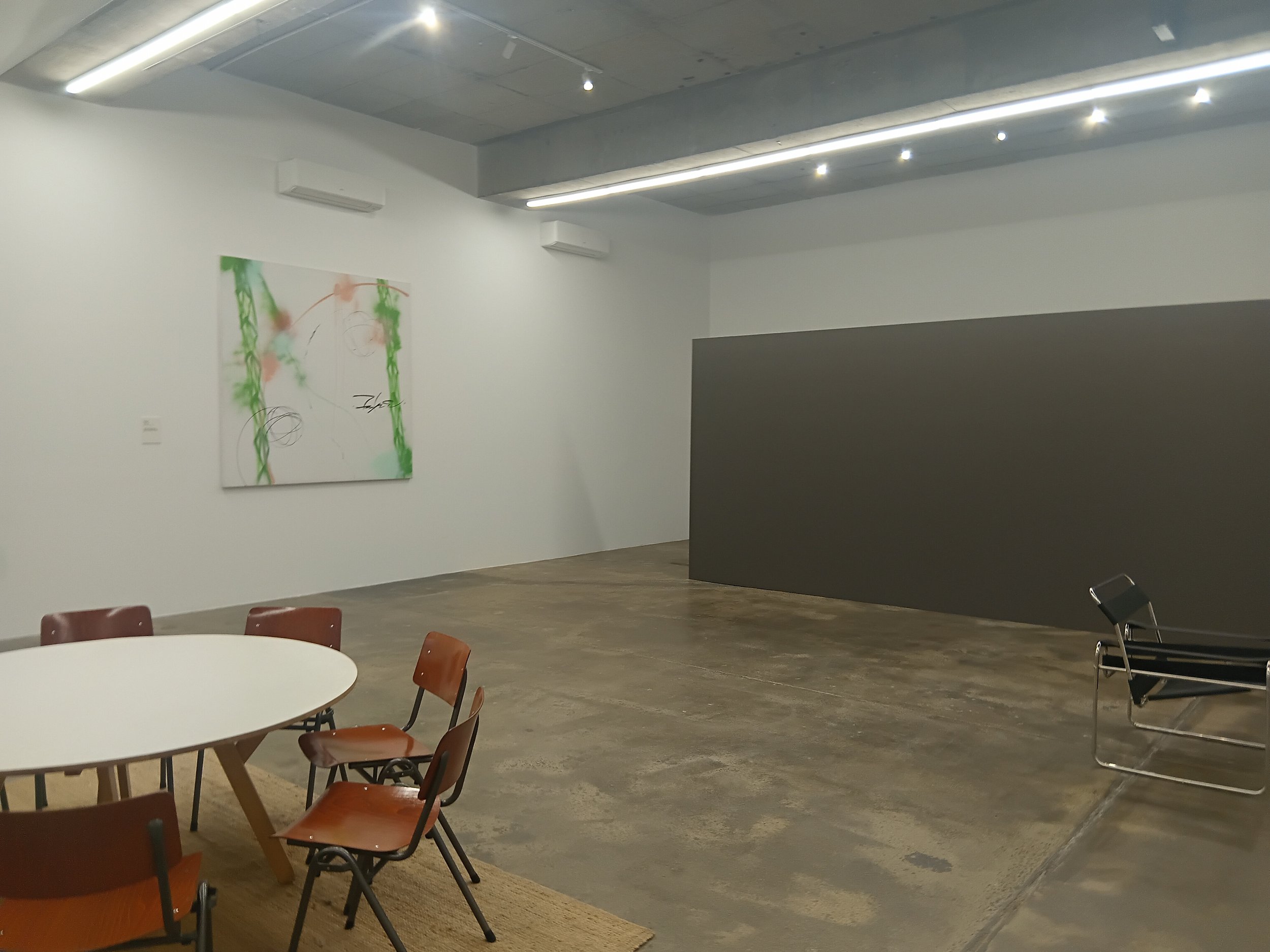 A modern art gallery or conference room with white walls, a concrete ceiling, and bright lighting. There is a colorful abstract painting on the wall, round tables with brown chairs, and a dark grey partial wall in the foreground. There are also chair