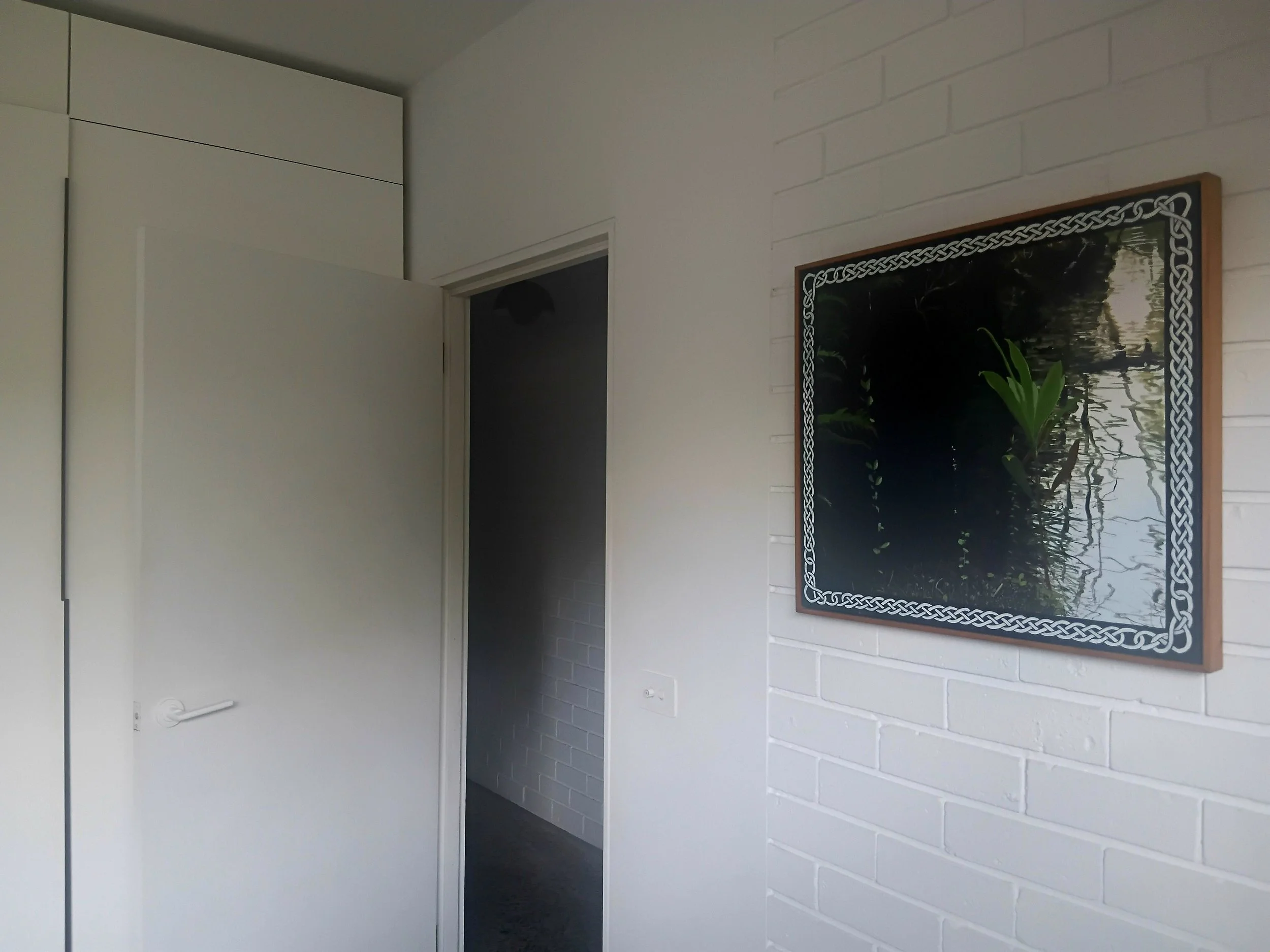 Interior view of a room with a white brick wall, a framed picture of a river scene, white cabinet, and a door opening to another room.