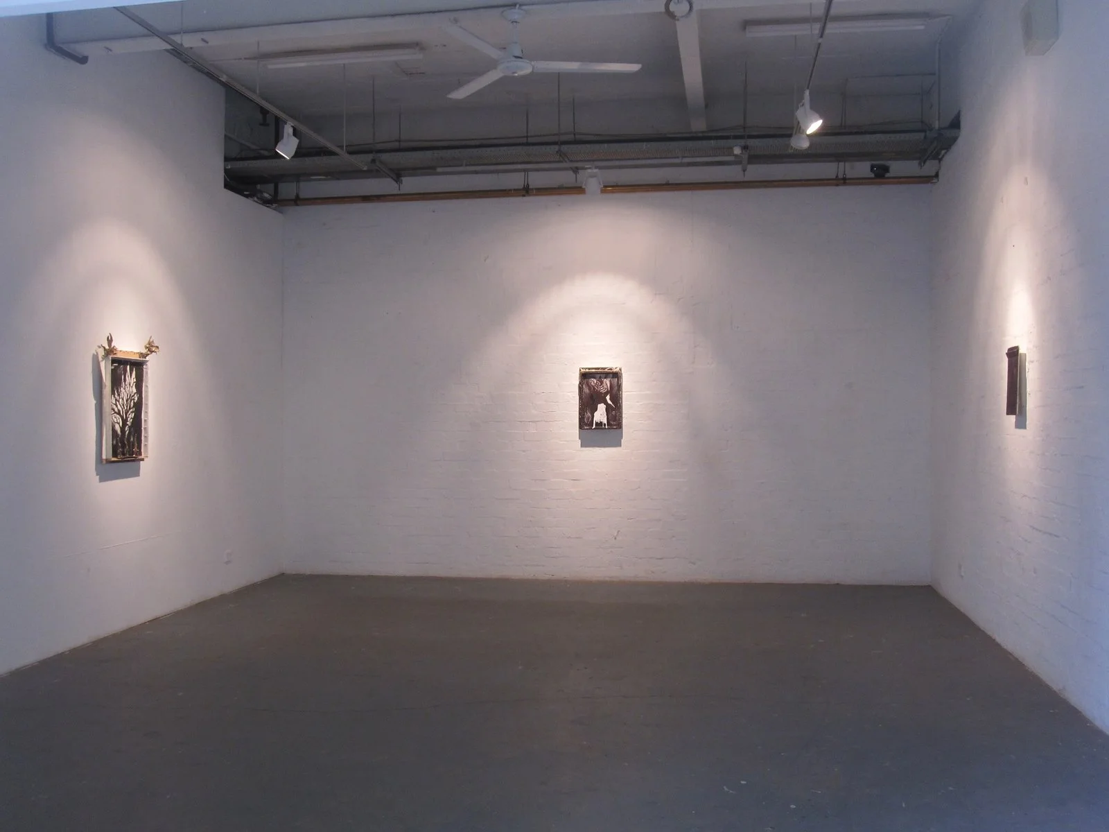 An art gallery with white brick walls, three small framed artworks, and track lighting.