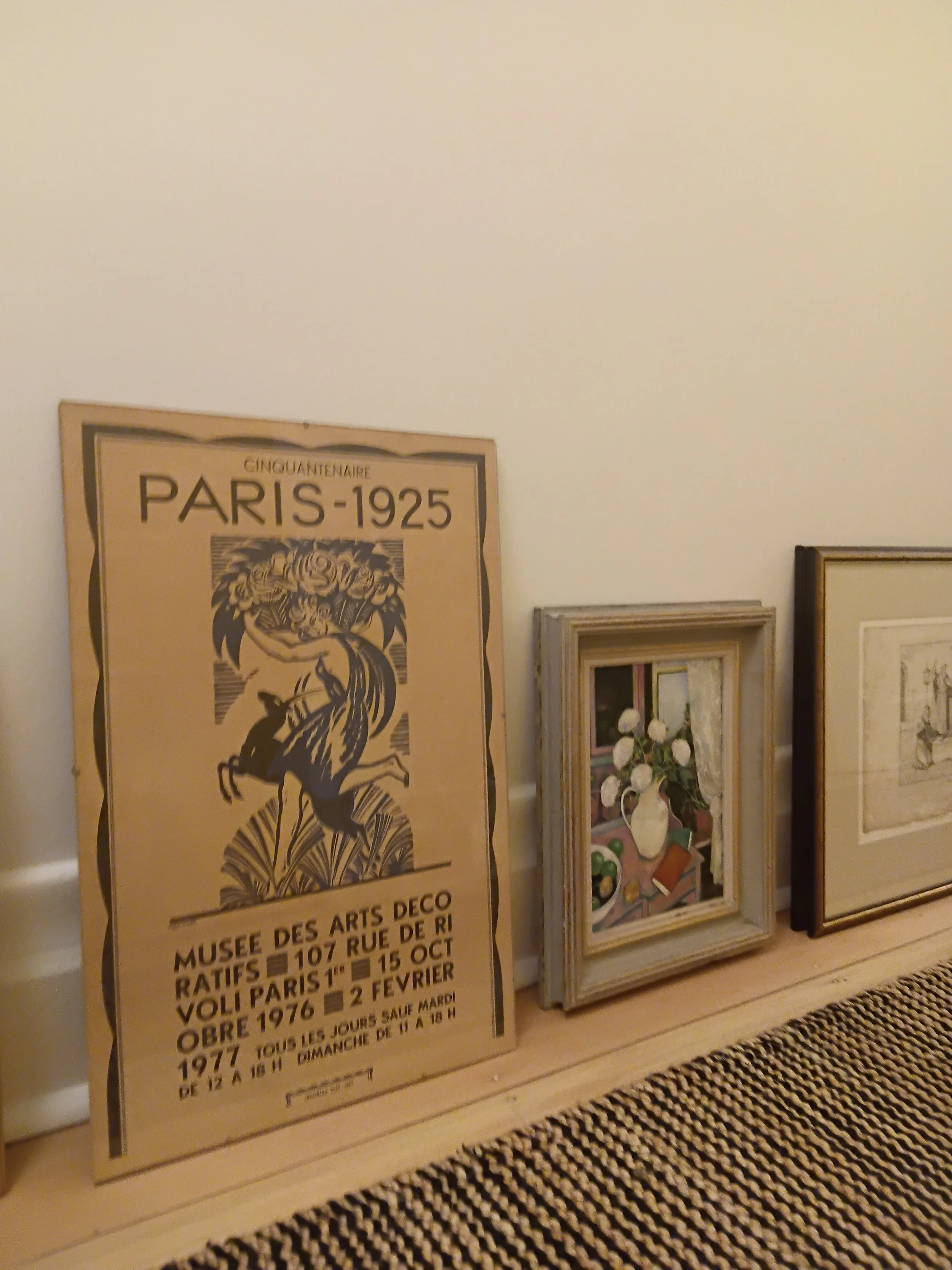 A large poster announcing Paris art exhibitions from 1925 to 1977, with text in French and some illustrations, leaning against a wall with framed pictures and artwork. Multiple frames and a floral painting are visible on the wall above.