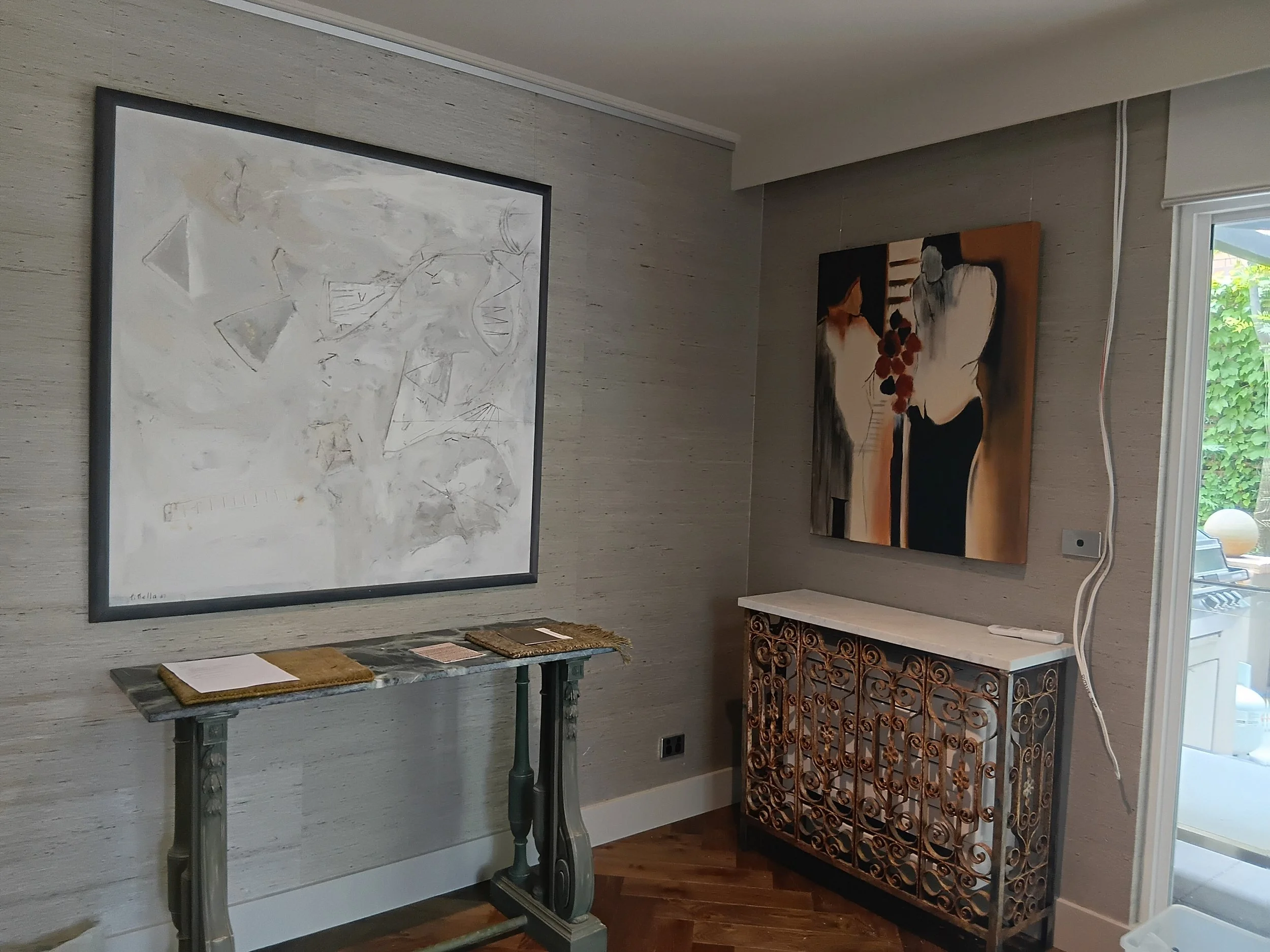 Interior of an art gallery with two abstract paintings on a wooden-textured wall, a marble-topped table with framed artwork and brown mats, and a decorative metal stand with intricate patterns next to a window with a view of green foliage.