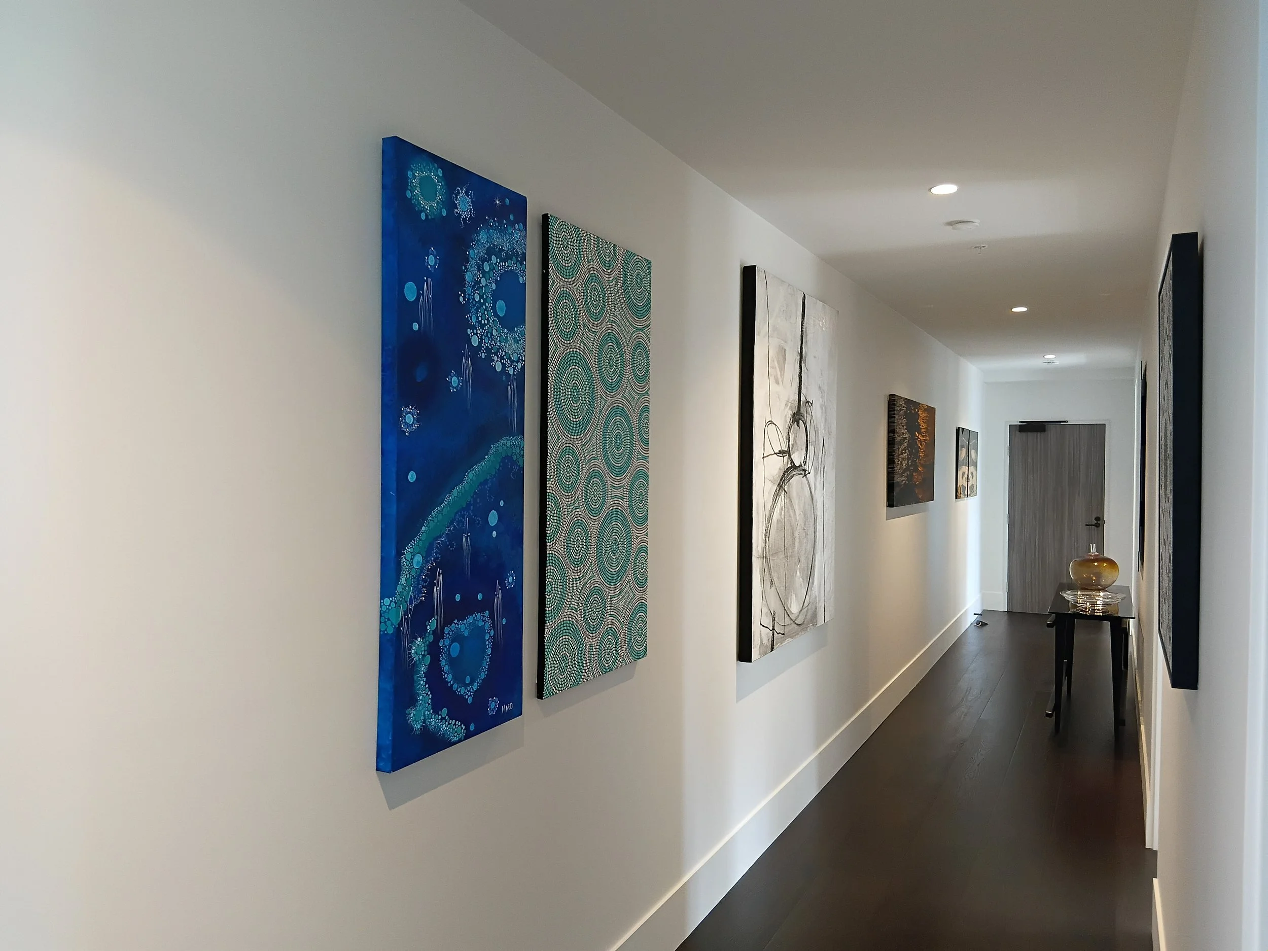 Image of a hallway with white walls decorated with various abstract paintings, and ceiling lights.