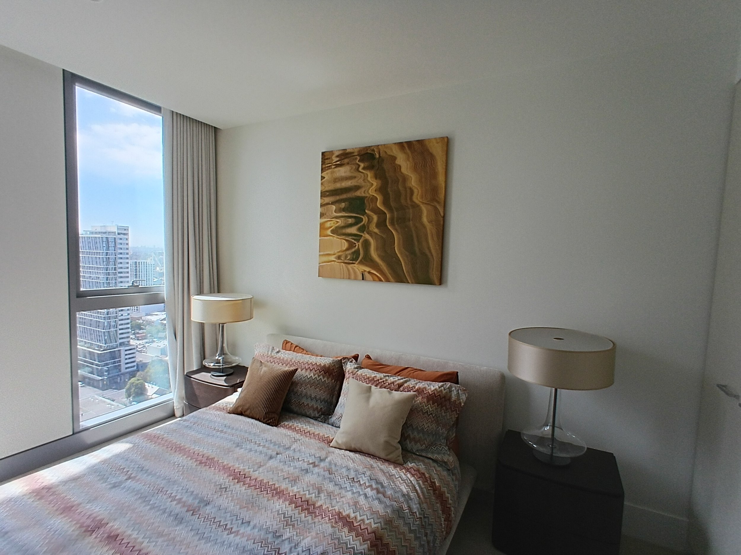 Modern bedroom with a bed covered in a zigzag patterned bedspread, several pillows, a nightstand with a lamp, a large window with city view, and a piece of abstract artwork on the wall.