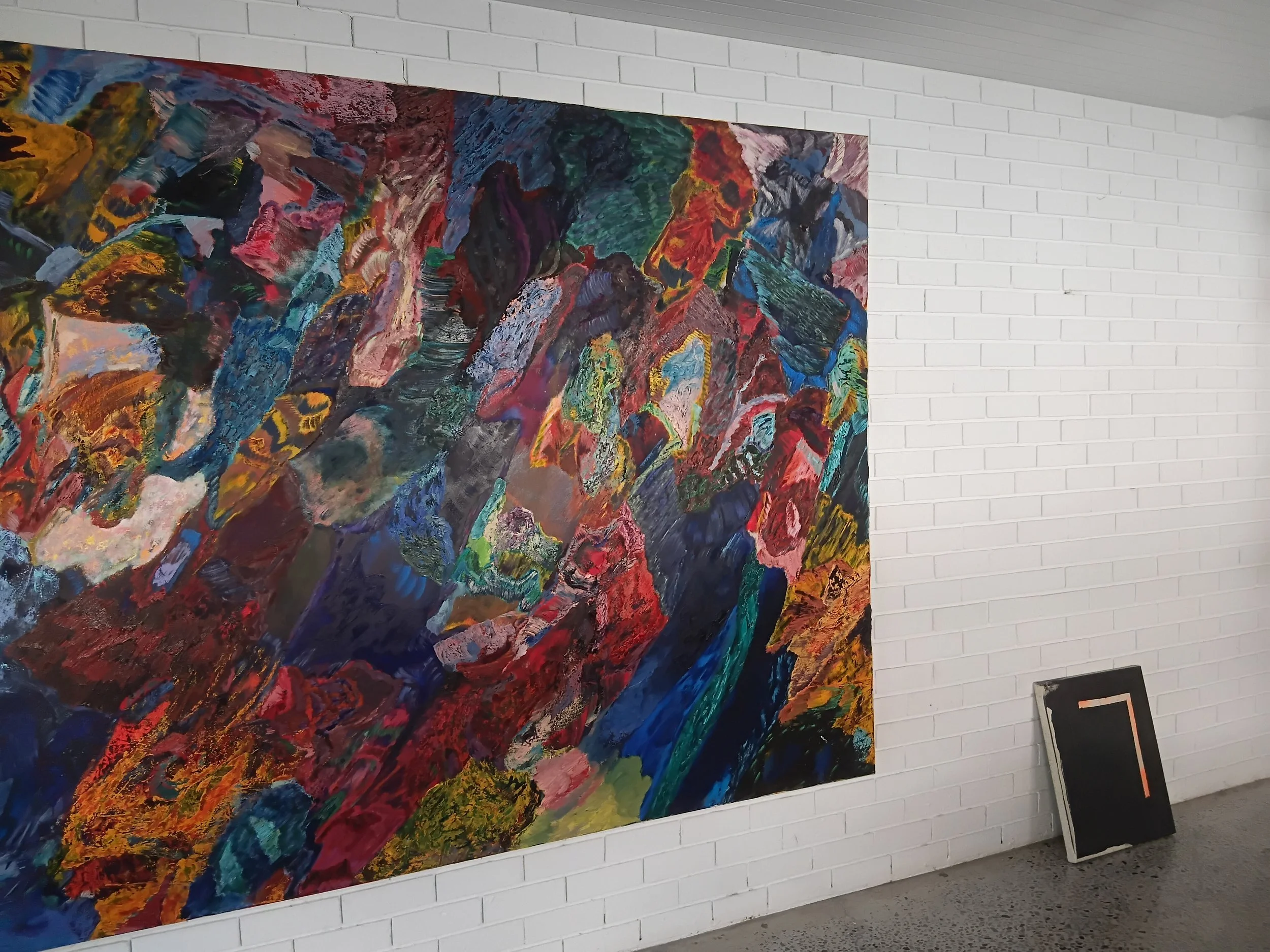 A colorful abstract painting displayed on a white brick wall in an indoor art gallery.