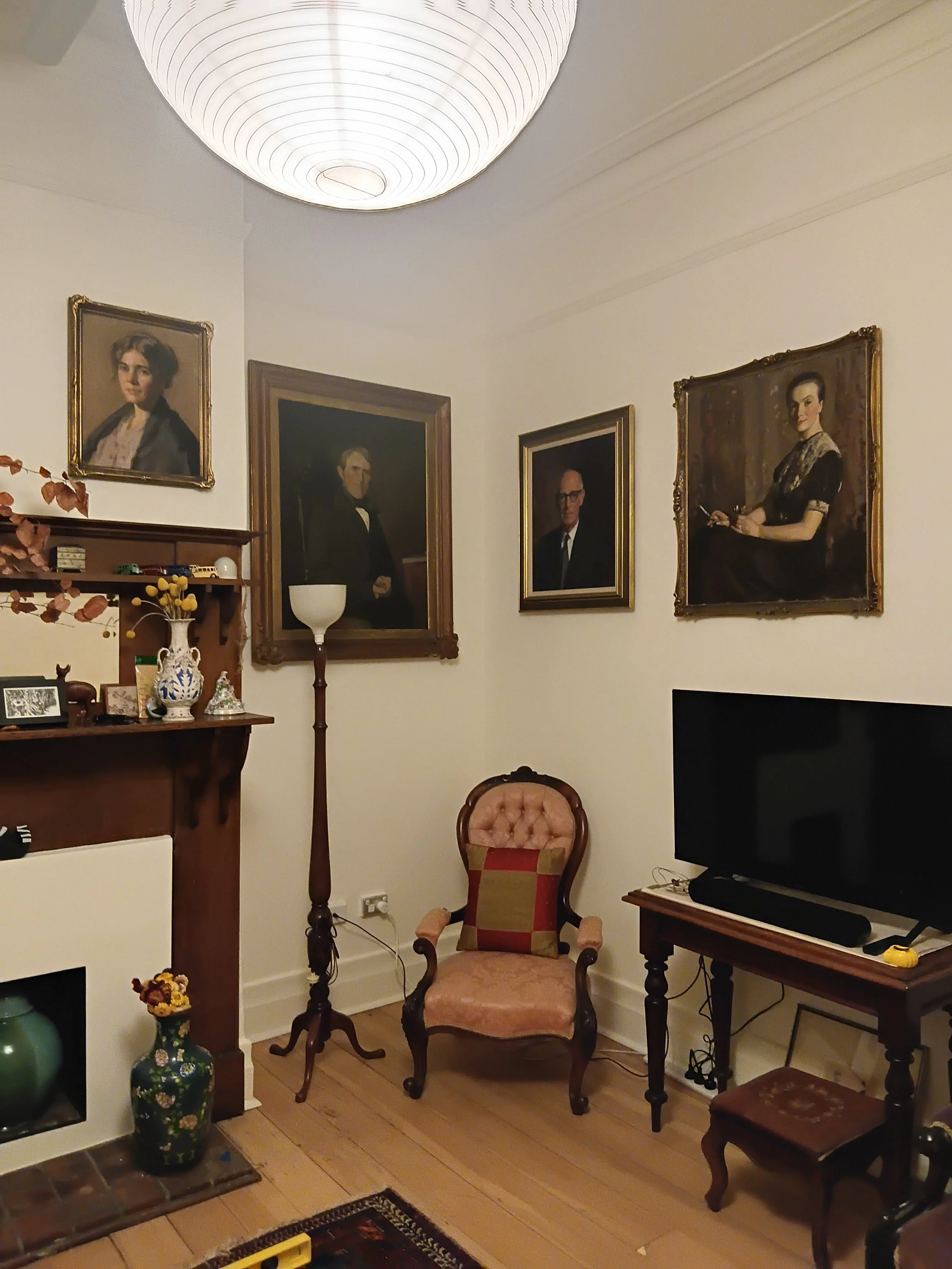 A cozy living room corner featuring vintage portraits of a man and a woman on the wall, a pink upholstered armchair, a small wooden table, a black television, and decorative items including a vase with flowers and small figurines. The room has a wood