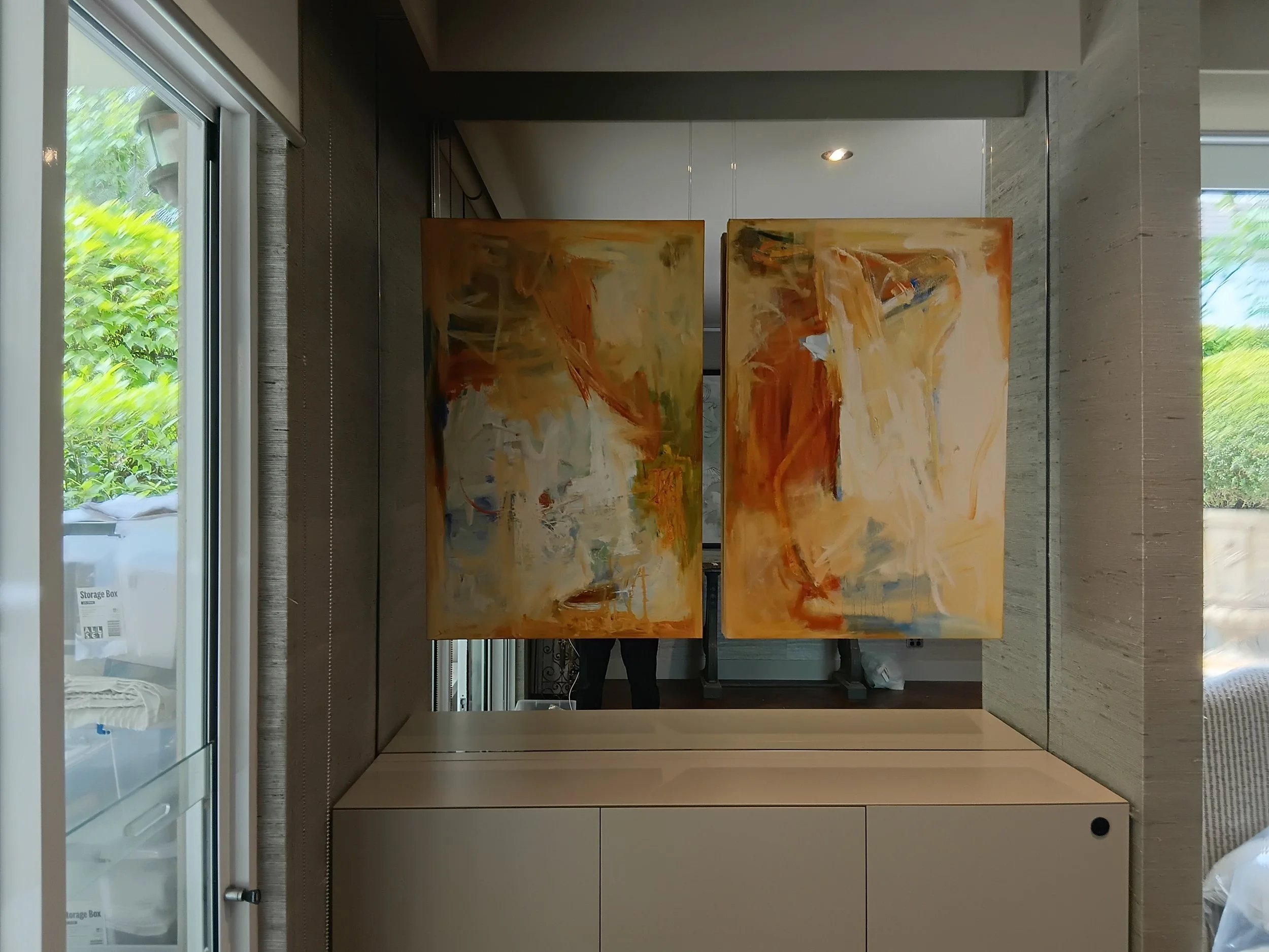Abstract painting with orange, beige, and white brushstrokes behind a mirror in a well-lit room with large windows and outdoor greenery.
