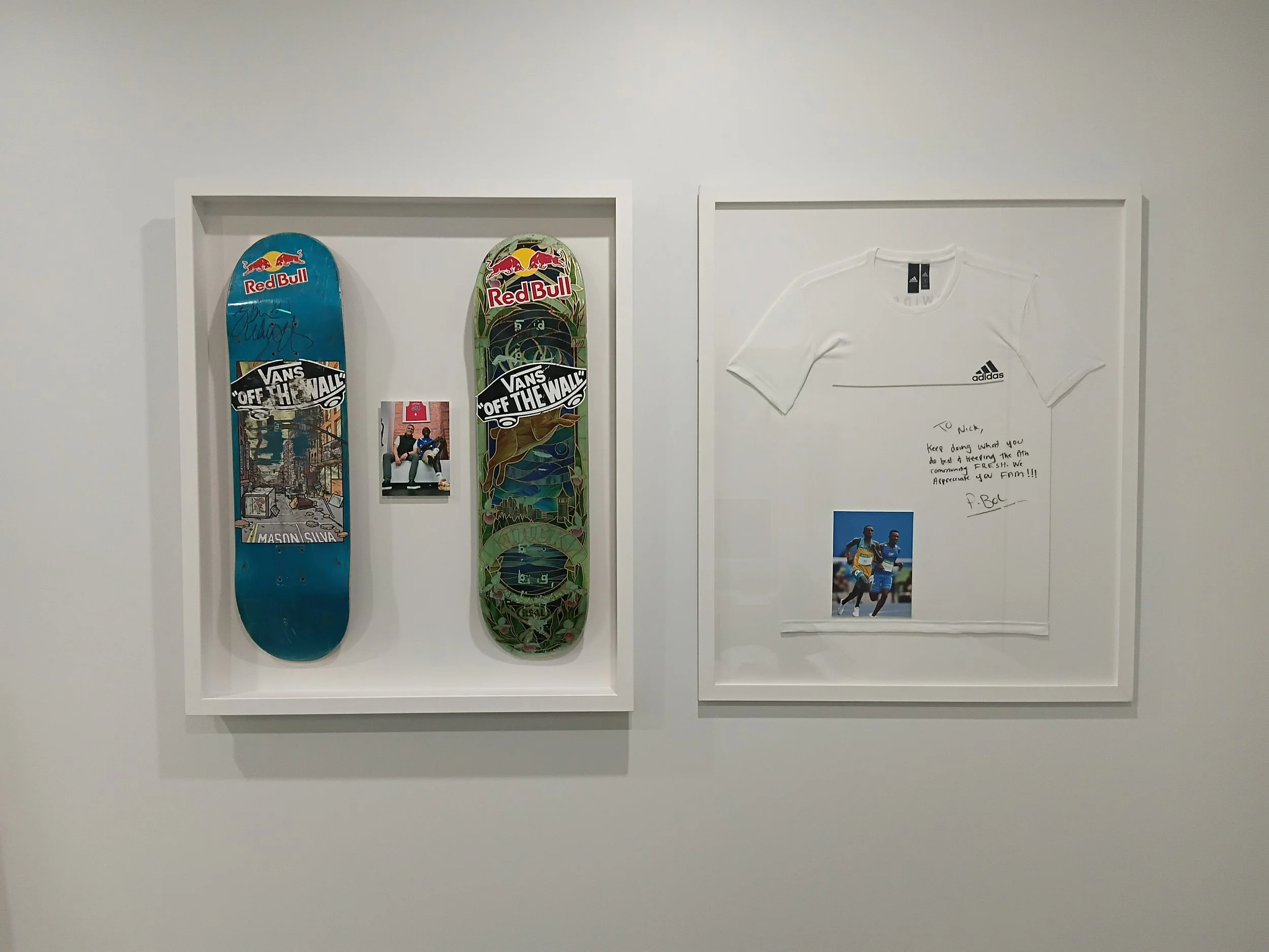 Two skateboards with Red Bull branding and a photograph of two runners, along with a white Adidas shirt with a handwritten note, framed and mounted on a white wall.
