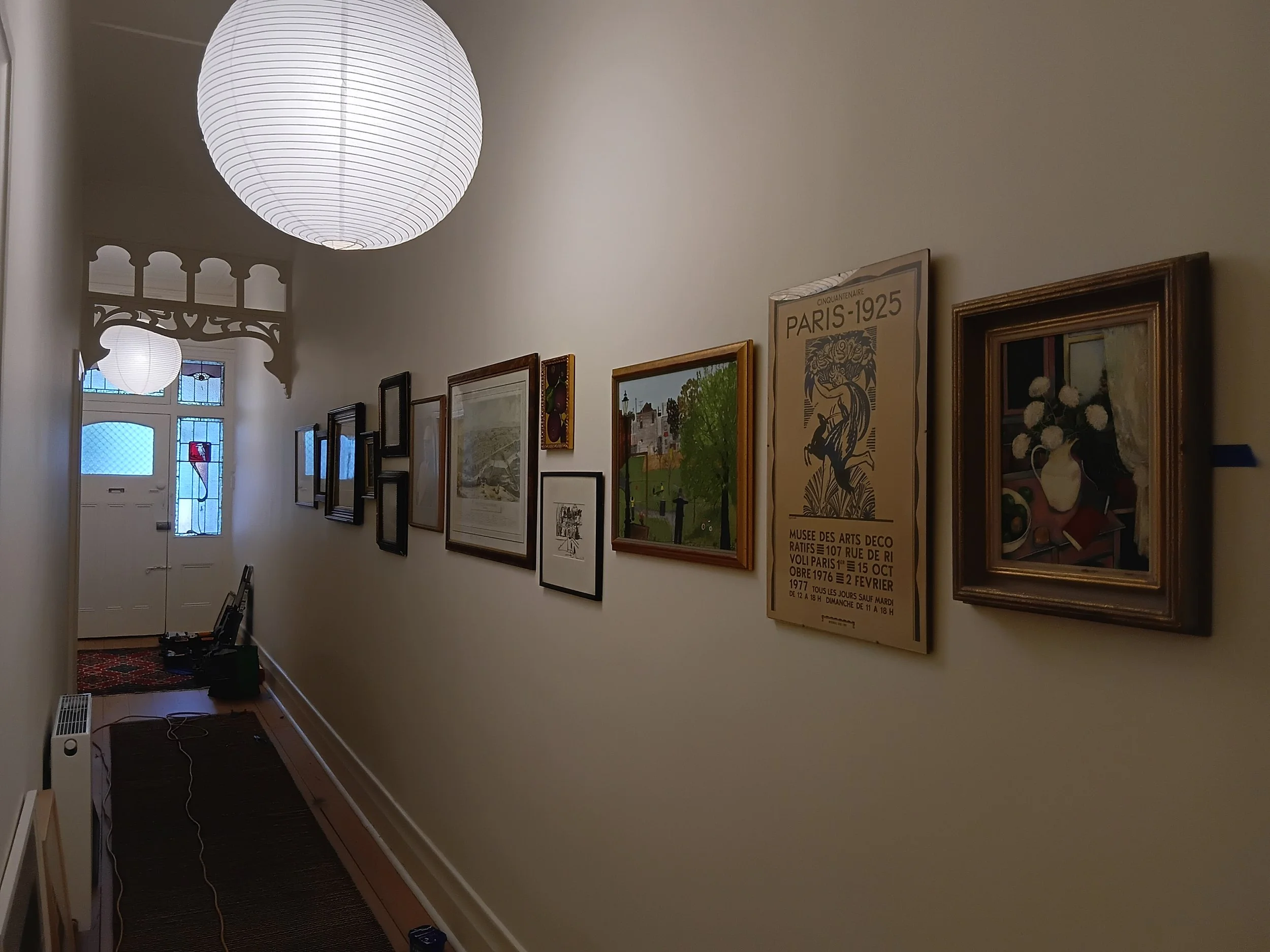 A hallway with framed artwork and posters on the wall, a large paper lantern ceiling light, and a window at the end of the hall with stained glass and a door below it.