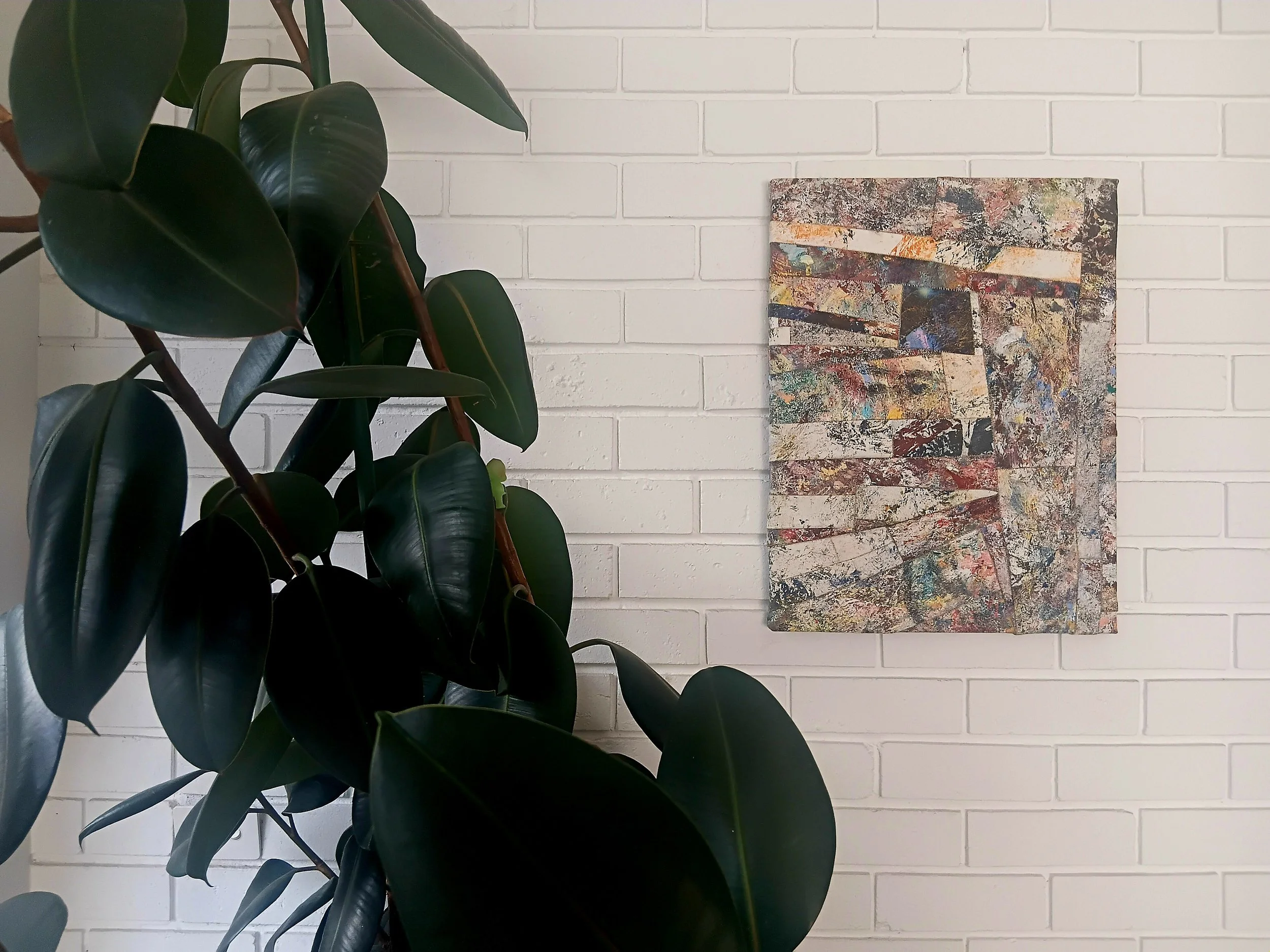 A close-up of a large houseplant with dark green, shiny leaves in front of a white brick wall, with a colorful abstract painting hanging on the wall.