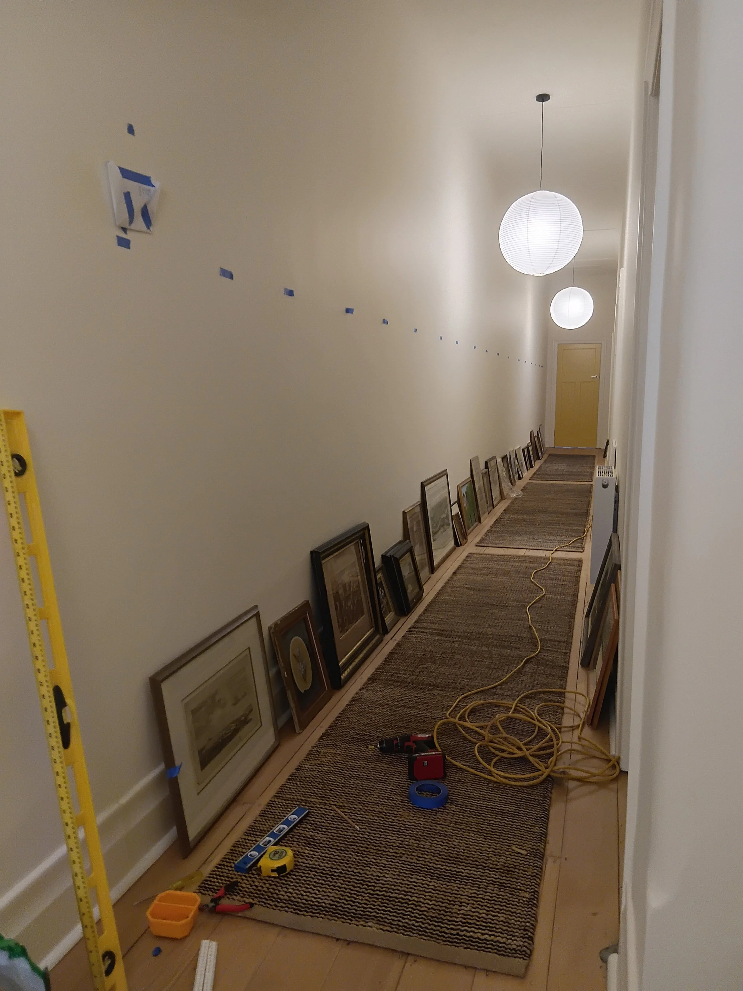 Hallway with framed pictures on the floor, yellow door at the end, three hanging paper lanterns, and tools and supplies on the floor.