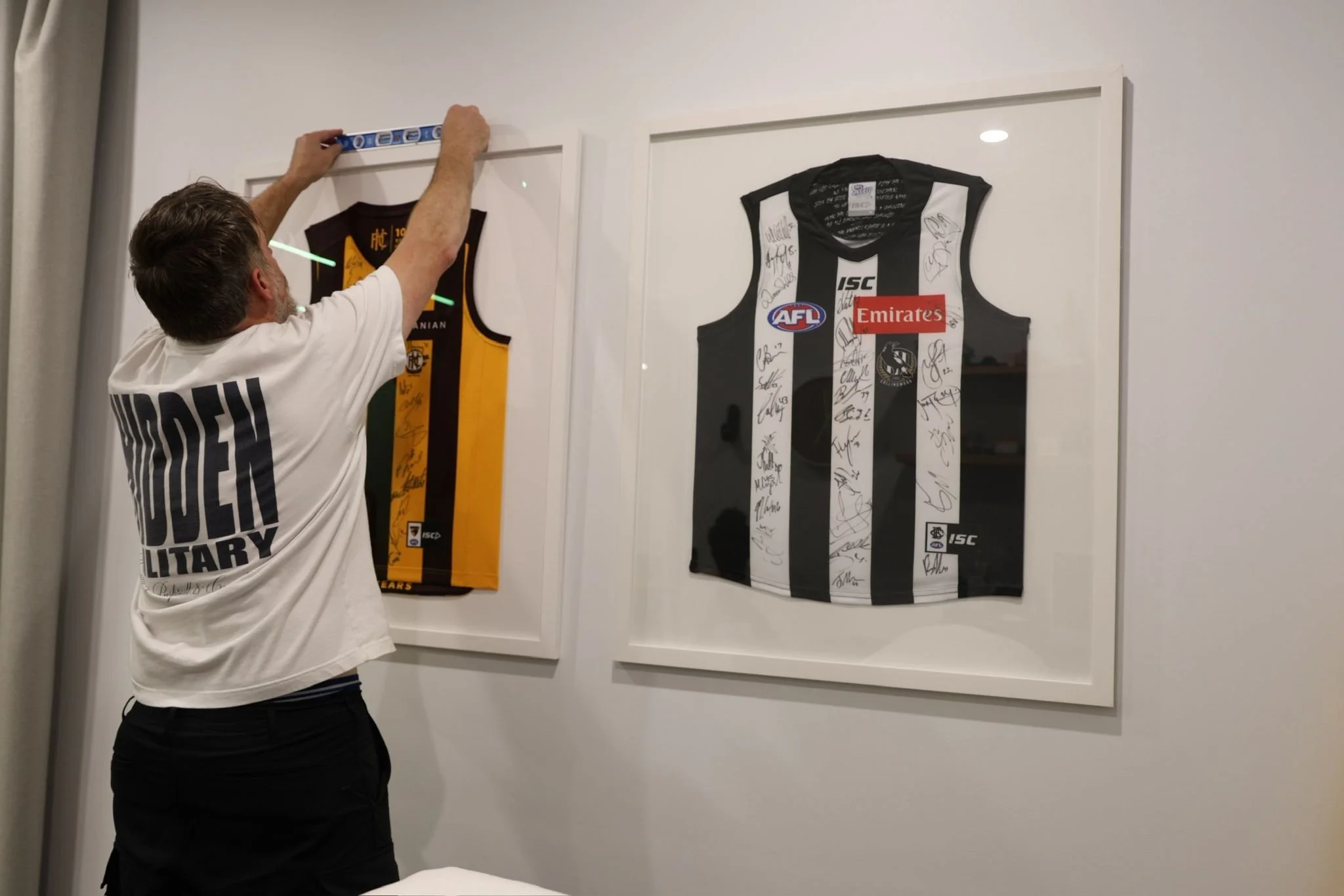 A man hanging a sports jersey on a wall, with two framed jerseys visible, one black and white signed jersey and a yellow and black framed jersey.