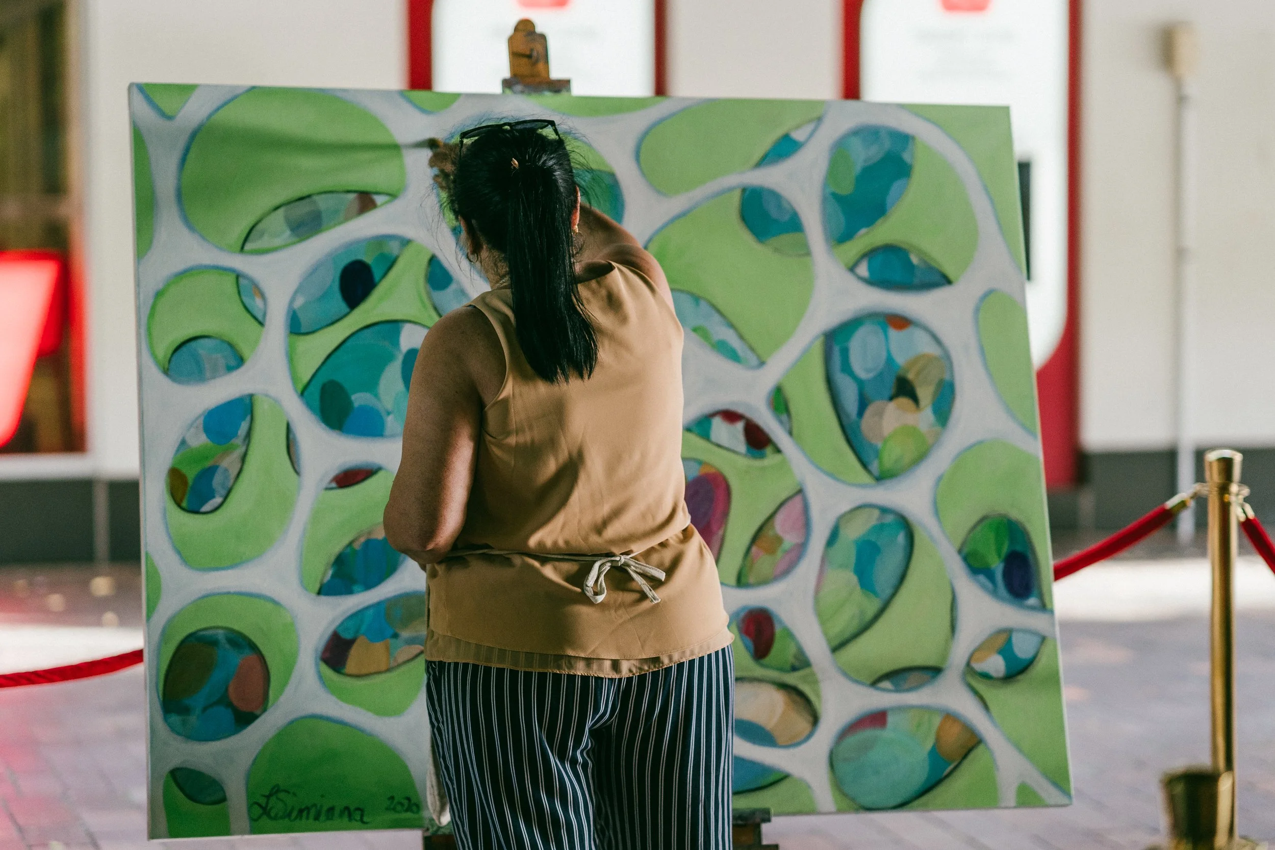 A woman with long dark hair tied in a ponytail, beige top, and striped pants, stands with her back to a colorful abstract painting with green, blue, and red shapes. The painting appears to be on an easel or display board. The setting is doors, possibly in an art exhibition or museum, with a red rope barrier visible in the foreground