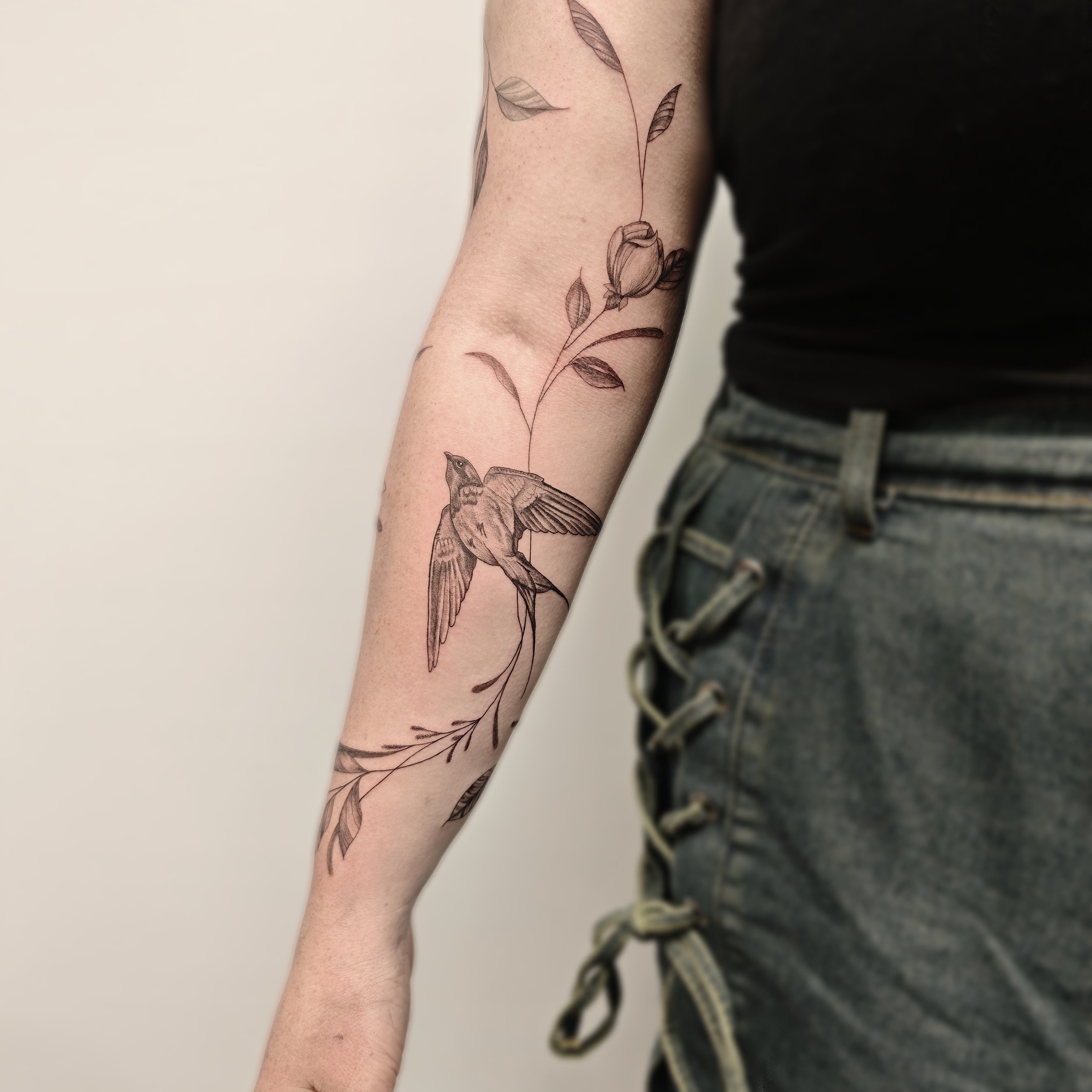 Tattoo am Unterarm mit einer Blume, einem Vogel und Blättern, alles in feiner Linienzeichnung.