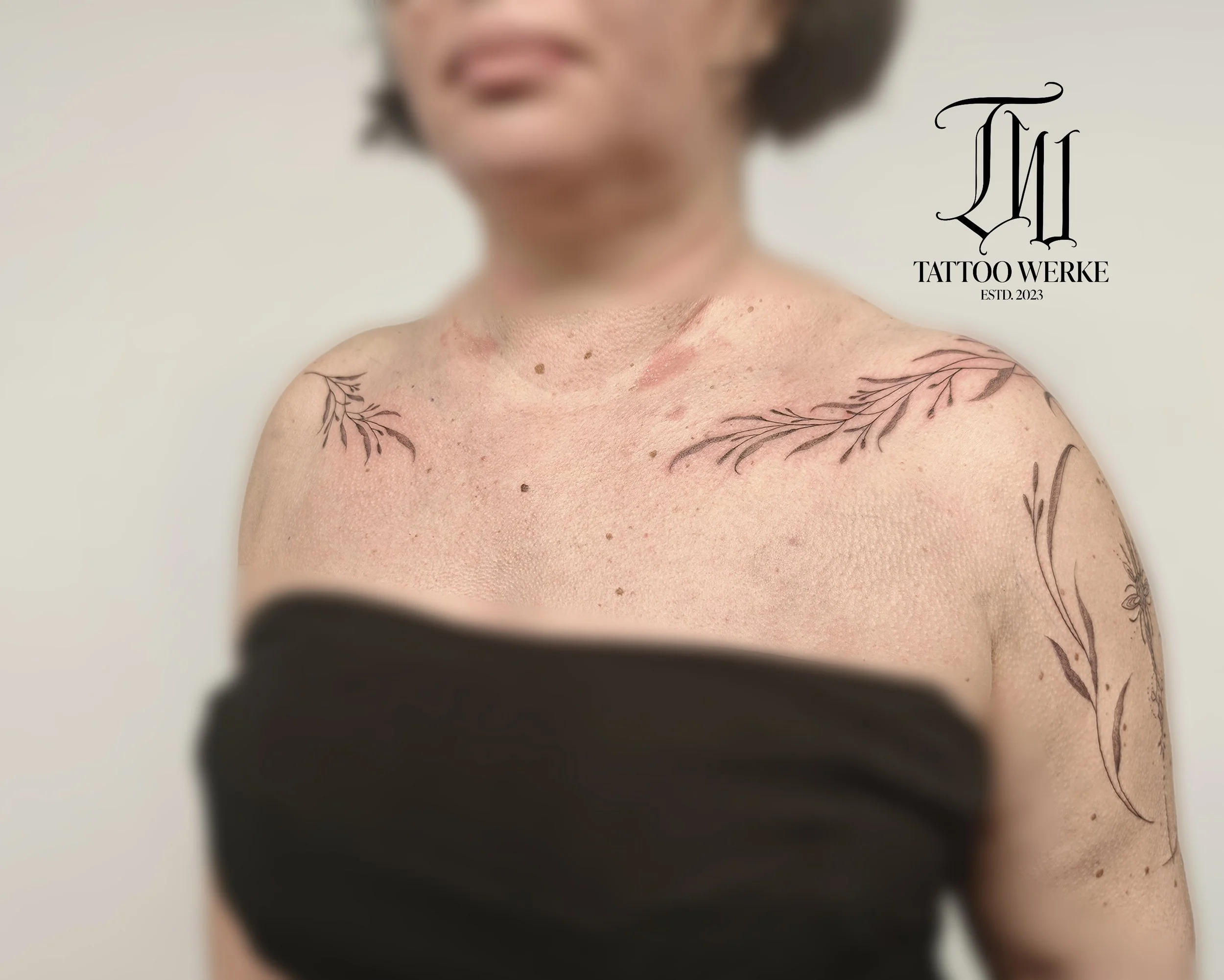 Naheaufnahme eines Schulterbereichs mit Tattoos auf beiden Schultern, eine ältere Frau mit kurzhaarigem Haar, unscharfer Kopf und Hals, weiße Wand im Hintergrund, Logo "Tattoo Werke" und Text "ESTD. 2023" rechts.