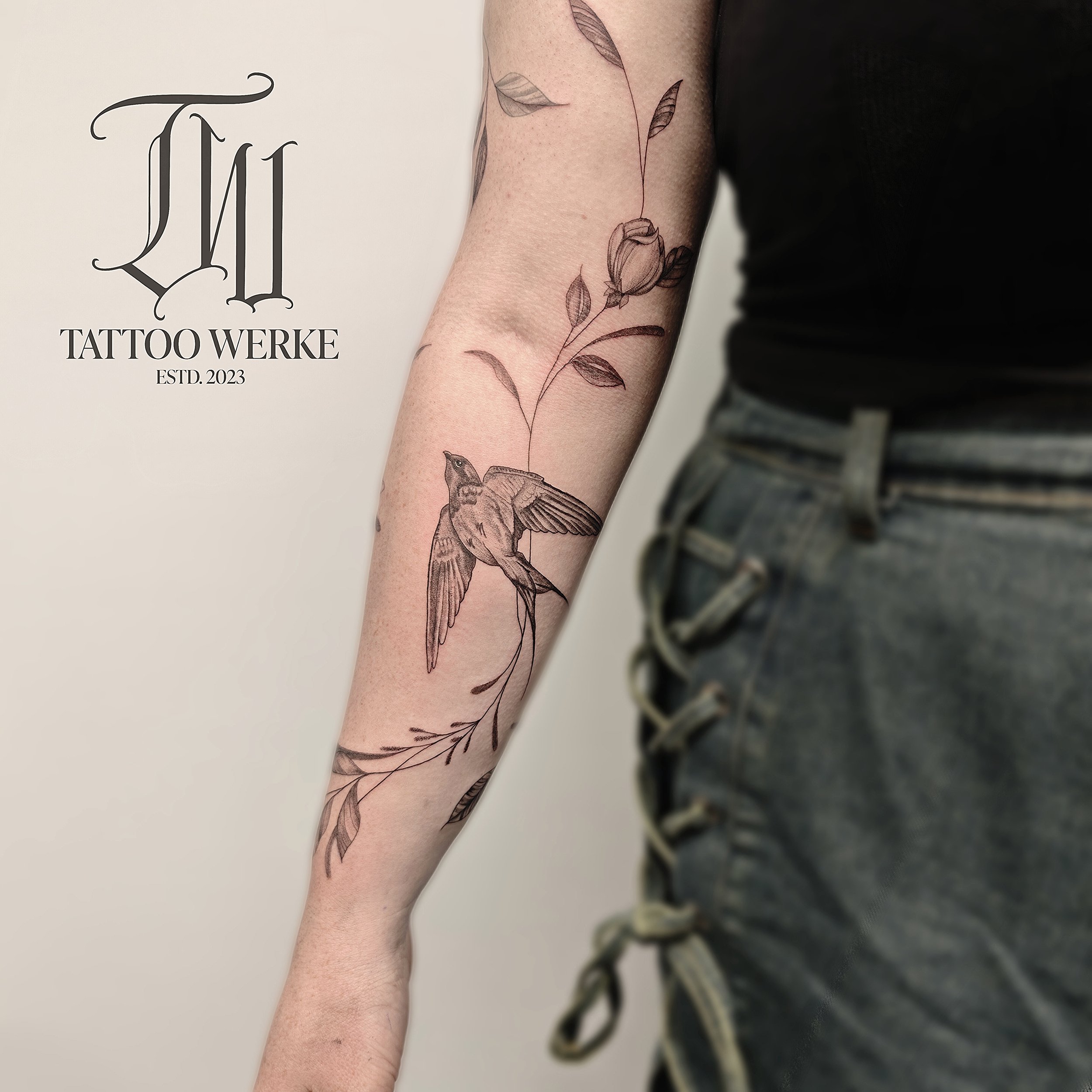 Eine Tätowierung auf einem Arm, die eine Blume, einen Vogel und einige Blätter in feinen Linien zeigt.