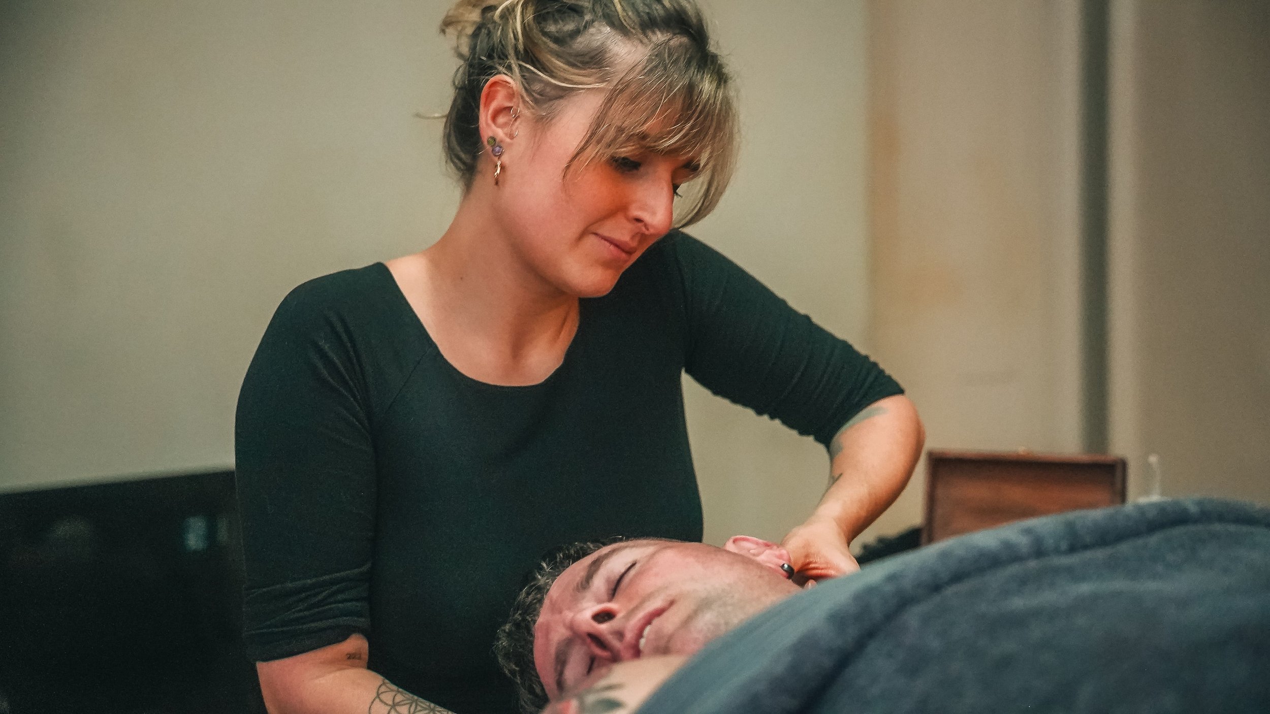 A woman with earrings and blonde hair styled in an updo is providing a massage or medical treatment to a man lying down with his eyes closed. The setting appears to be indoors, possibly in a clinic or home.
