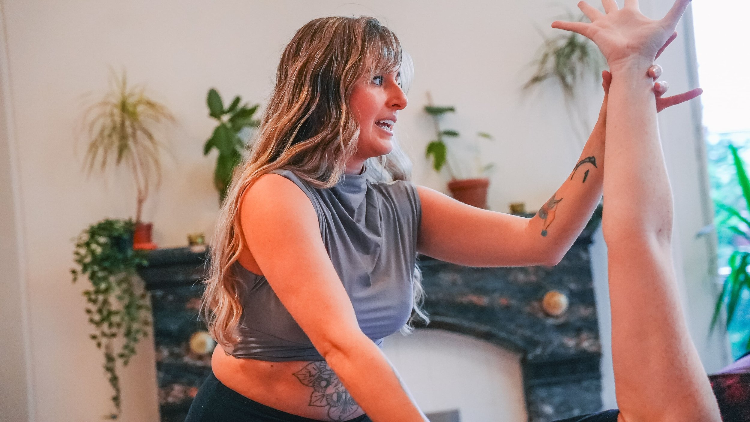 A woman with tattoos on her arm and waist is practicing a yoga pose indoors, with plants and furniture in the background.