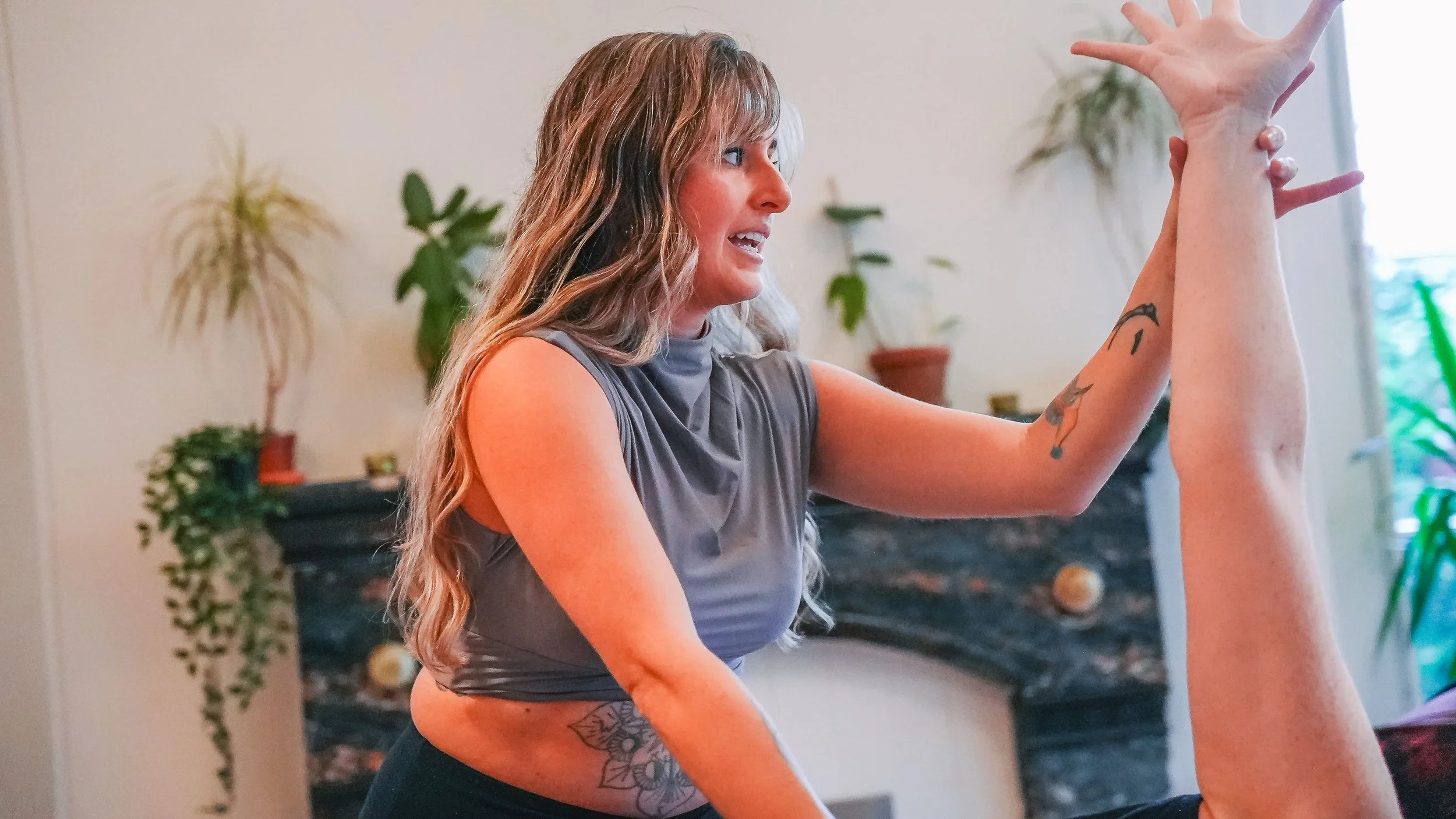 A woman with long, wavy blonde hair gesturing with her right hand during a conversation or activity indoors, with plants and a fireplace in the background.