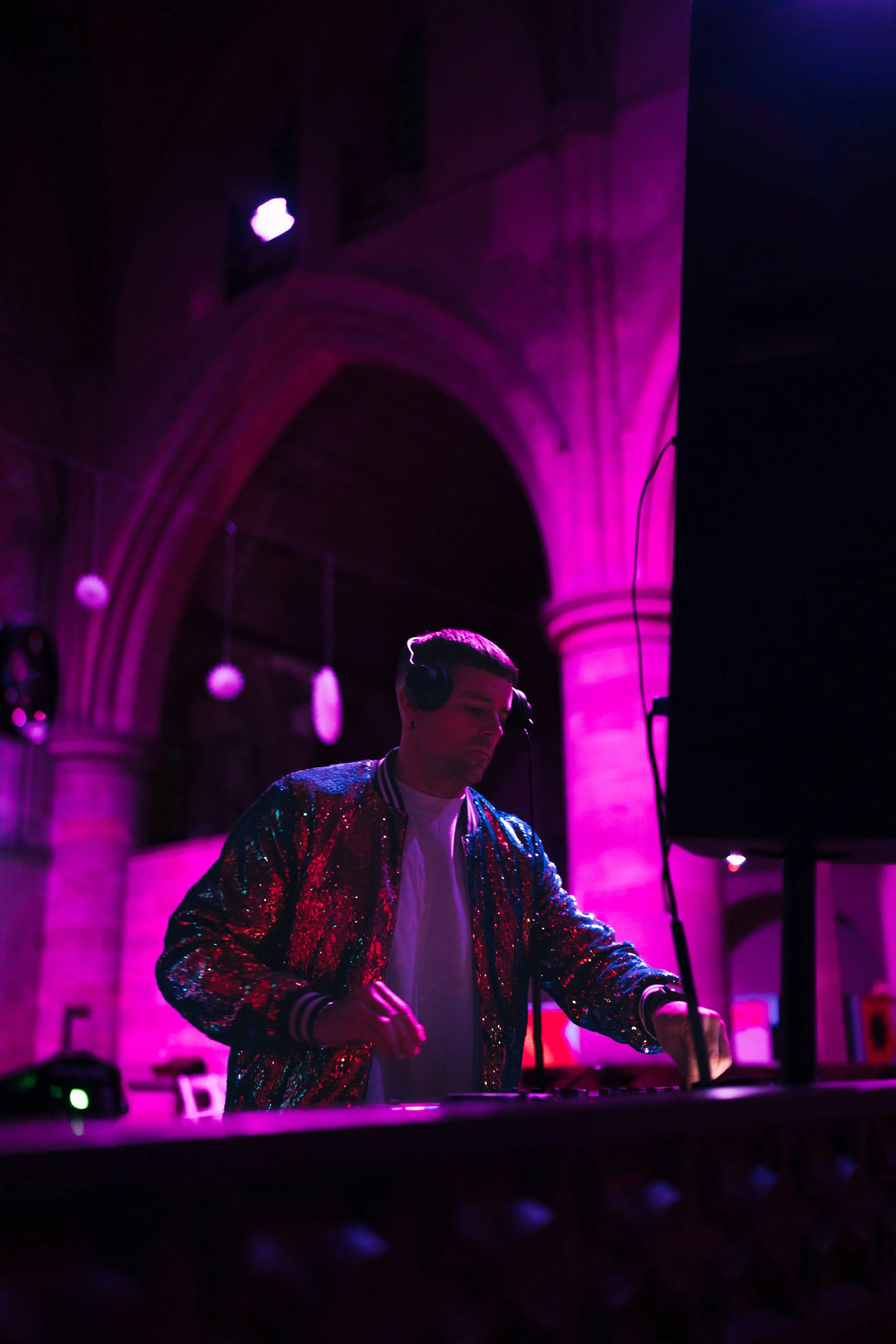 A DJ wearing a shiny, multicolored jacket is performing in a venue with pink/purple lighting and arched architecture.