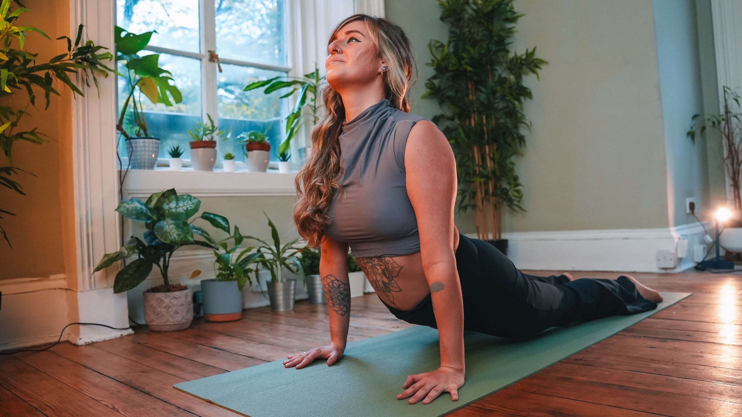 A woman practicing yoga on a green mat indoors, surrounded by potted plants near a window, with sunlight coming through.