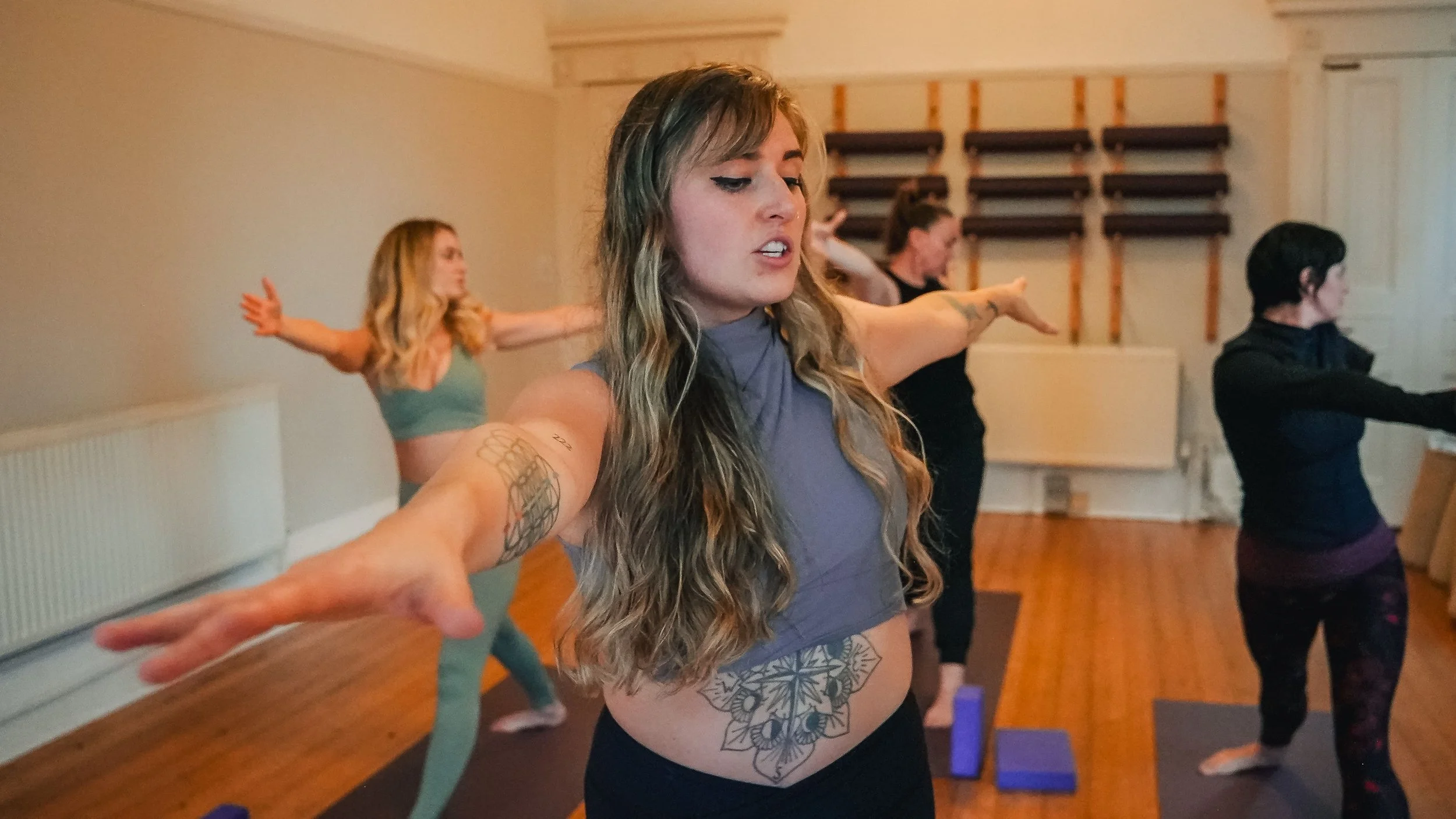A group of women practicing yoga in a studio, with one woman in the foreground focusing and extending her arms.