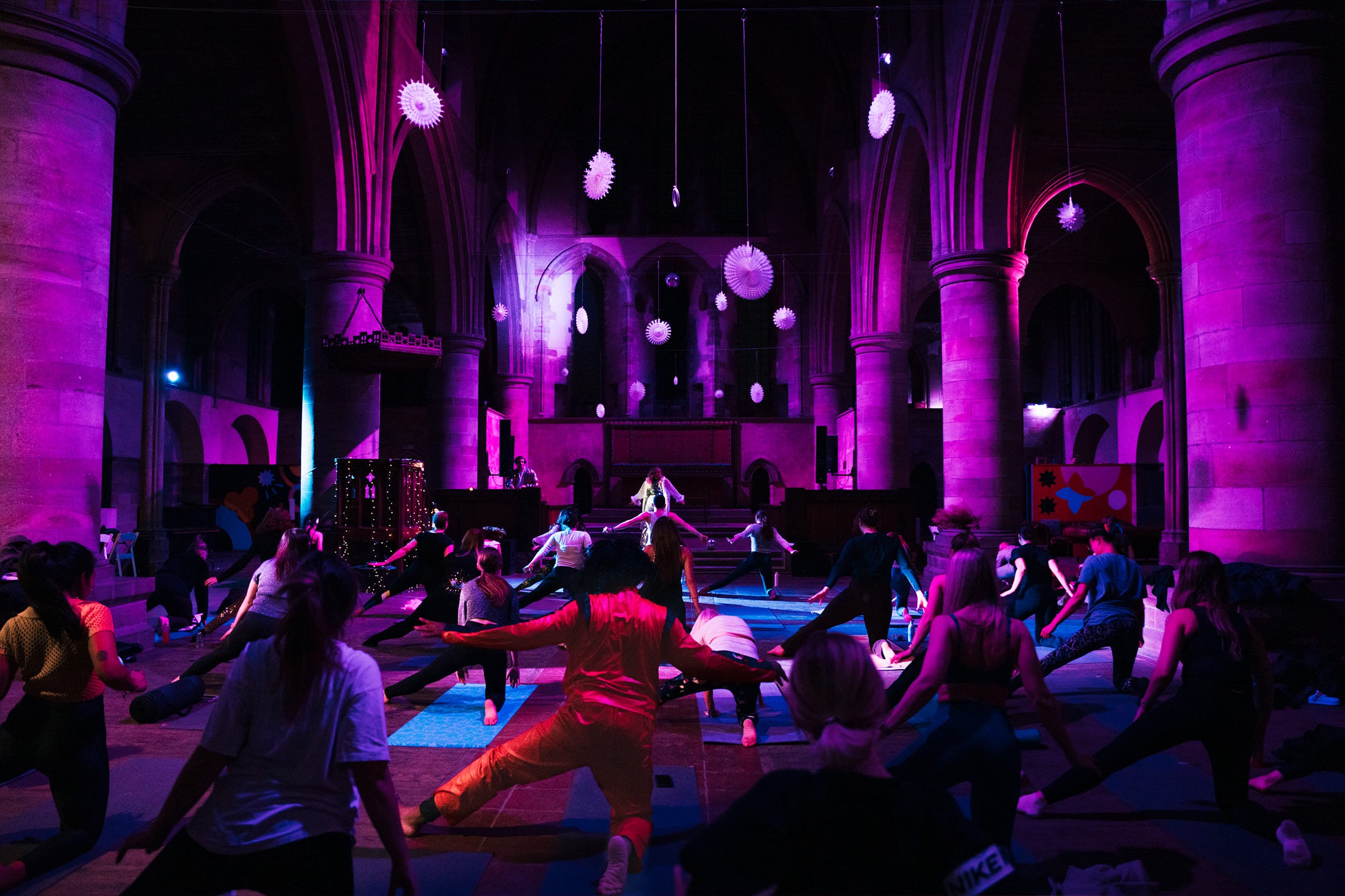 People participating in a yoga or fitness class inside a gothic cathedral lit with purple and pink lights, with hanging decorations