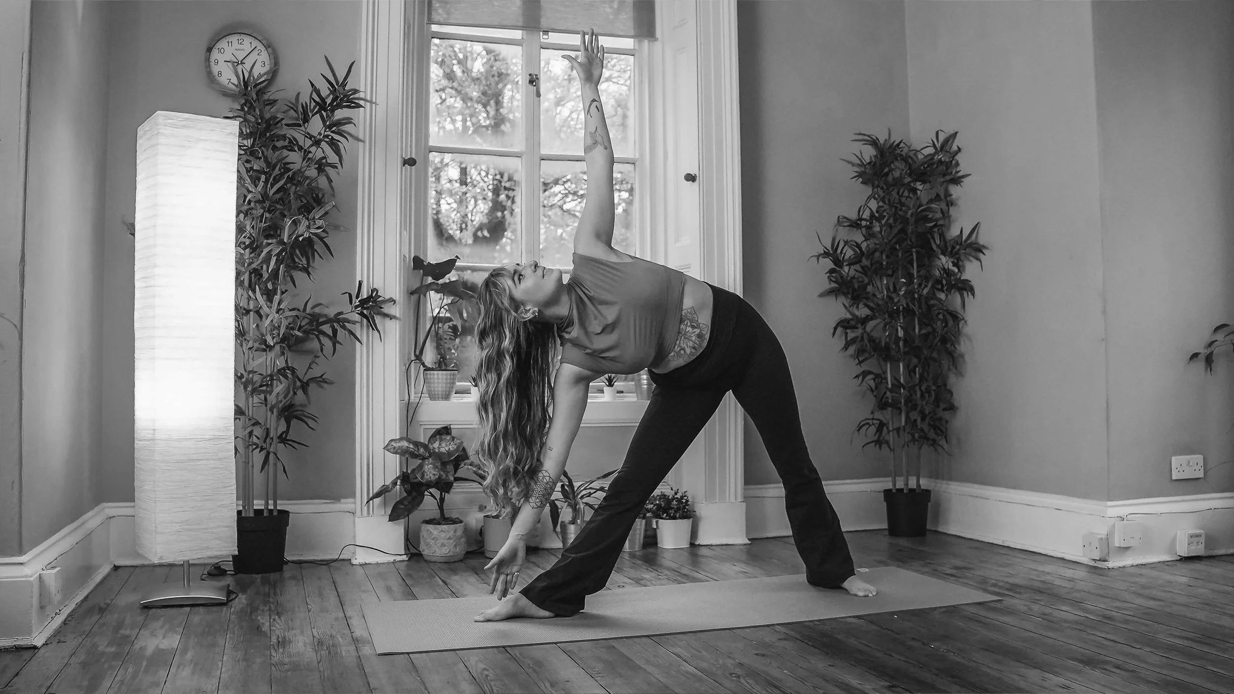 A woman practicing yoga indoors. She is in a side bend pose on a mat near a window, with planters and a tall floor lamp in the background.
