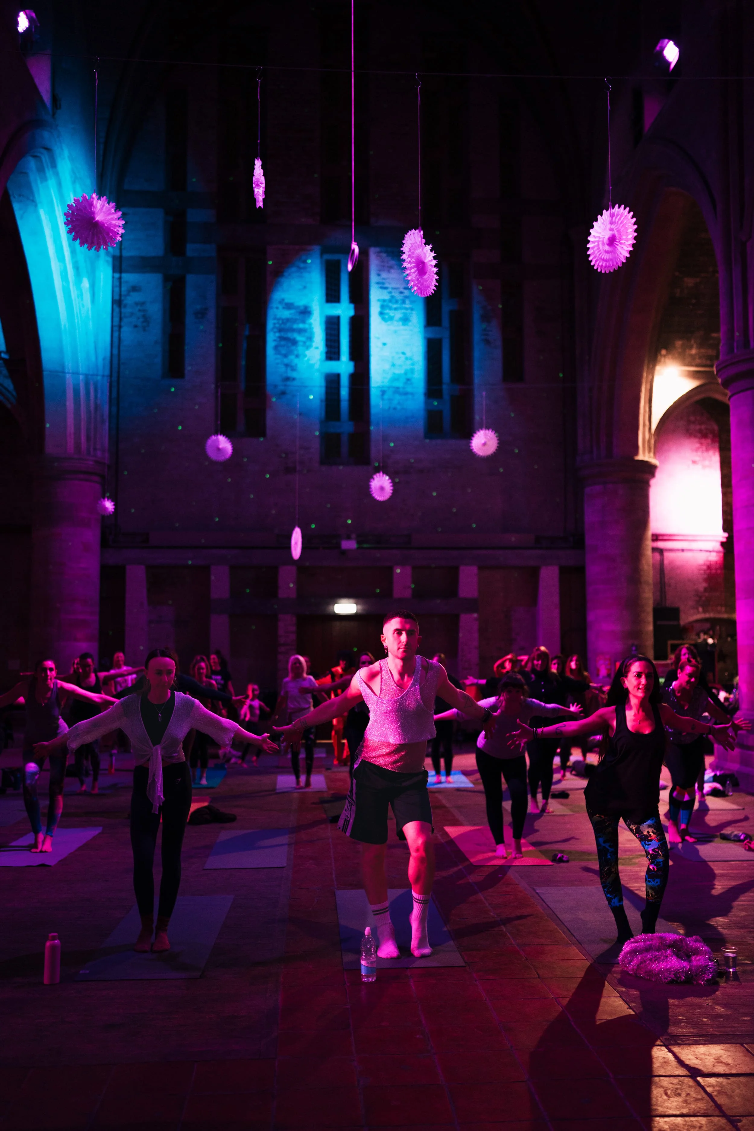 A group of people participating in a yoga class or dance class inside a large, dimly lit hall. The ceiling is decorated with pink and purple hanging paper decorations. The hall features high, arched ceilings and stained glass windows, with colored lighting casting pink, purple, and blue hues across the space. Participants are standing on yoga mats, with a woman at the front wearing a shiny top and shorts, surrounded by others in various workout outfits.