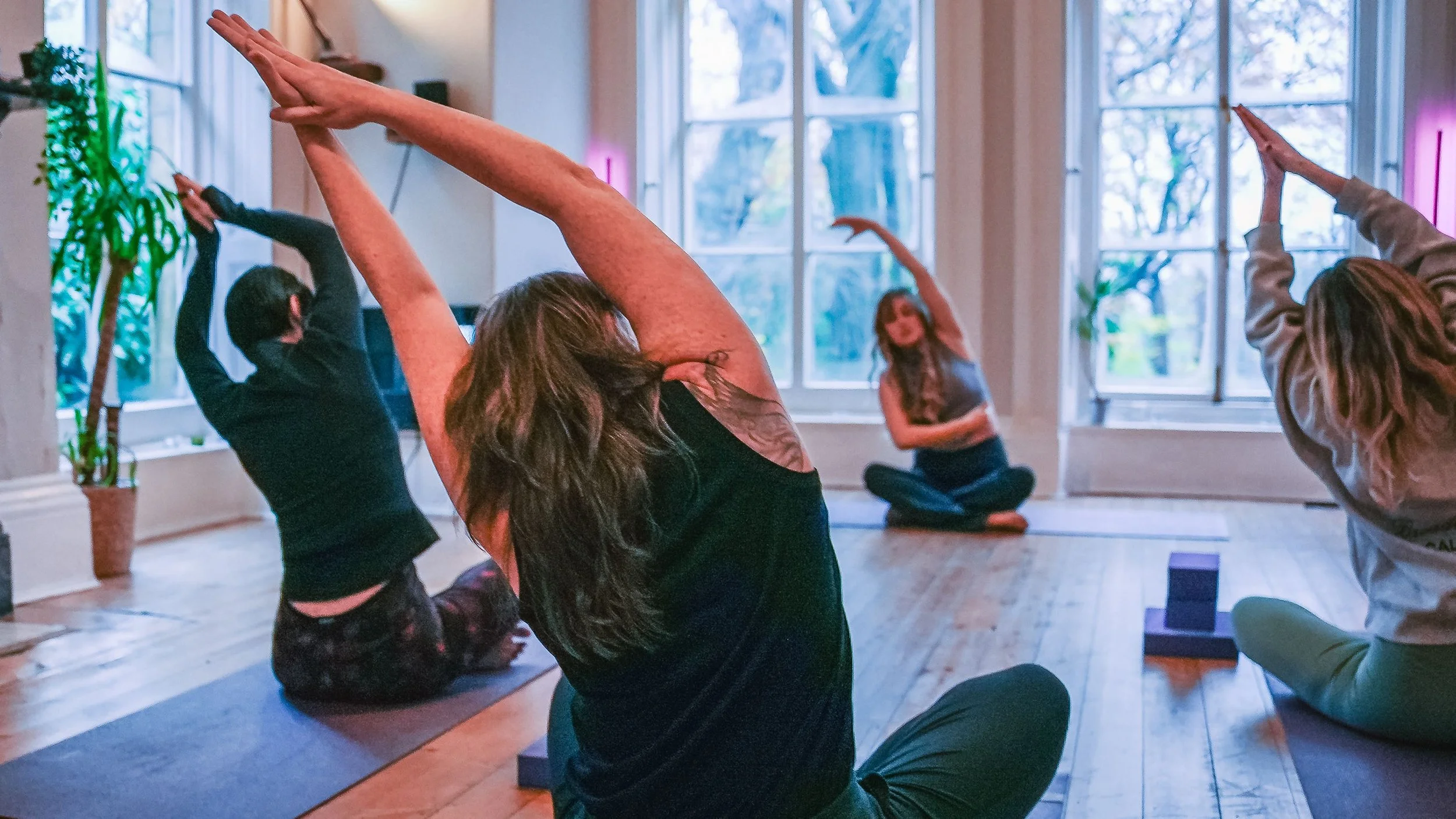People doing yoga or meditation in a bright indoor space with large windows, some sitting on mats and others stretching.