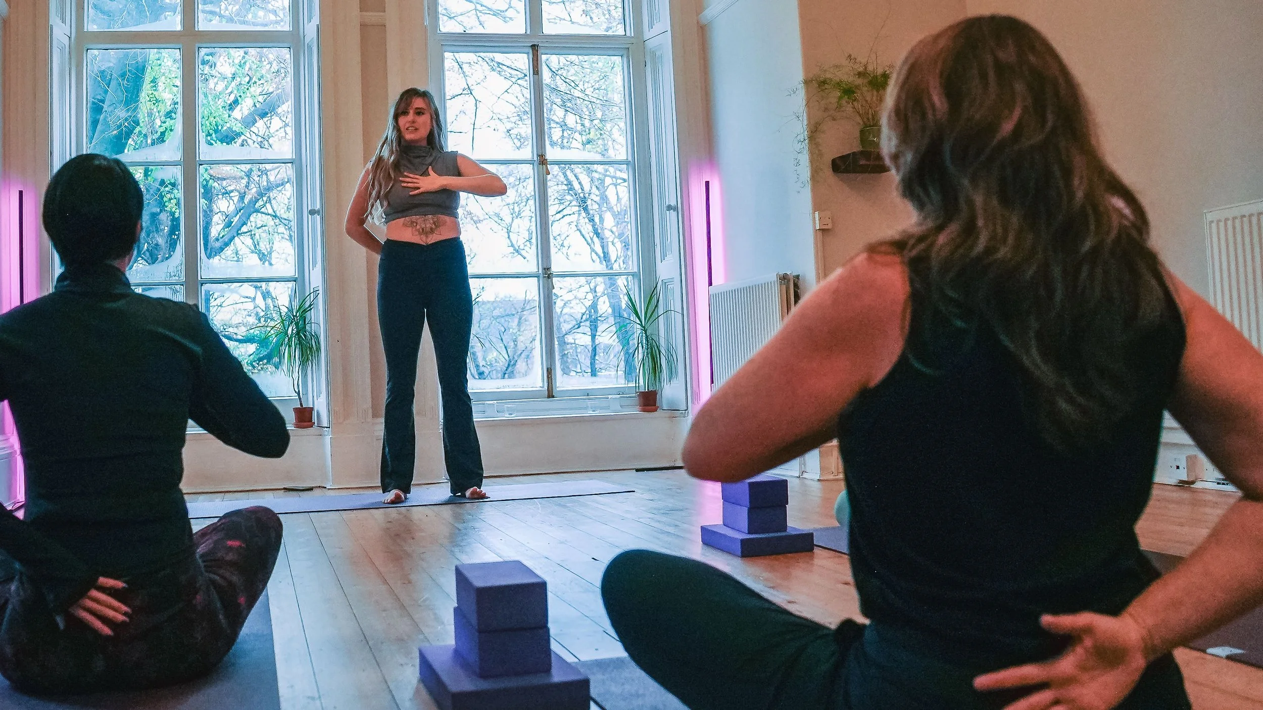 A woman leading a yoga or meditation class in a bright room with large windows, with two students seated on mats in front, one with black hair and another with brown hair, and yoga blocks and potted plants nearby.