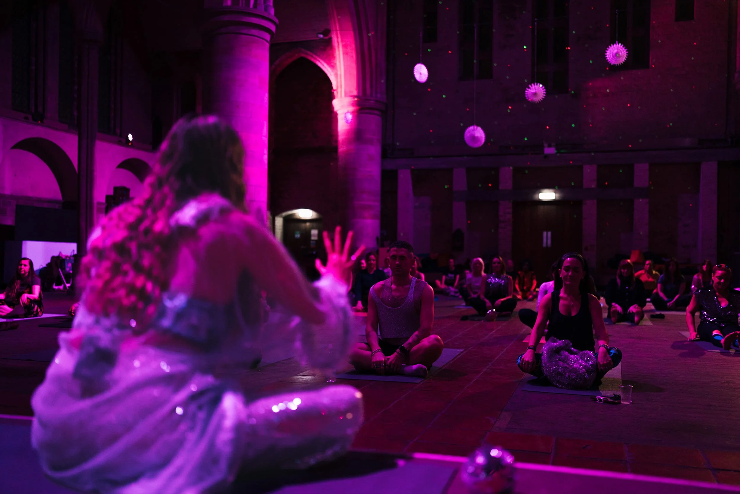 People sitting cross-legged on the floor in a dimly lit room with purple and pink lighting, participating in a group meditation or yoga session; decorations resembling virus particles are hanging from the ceiling.