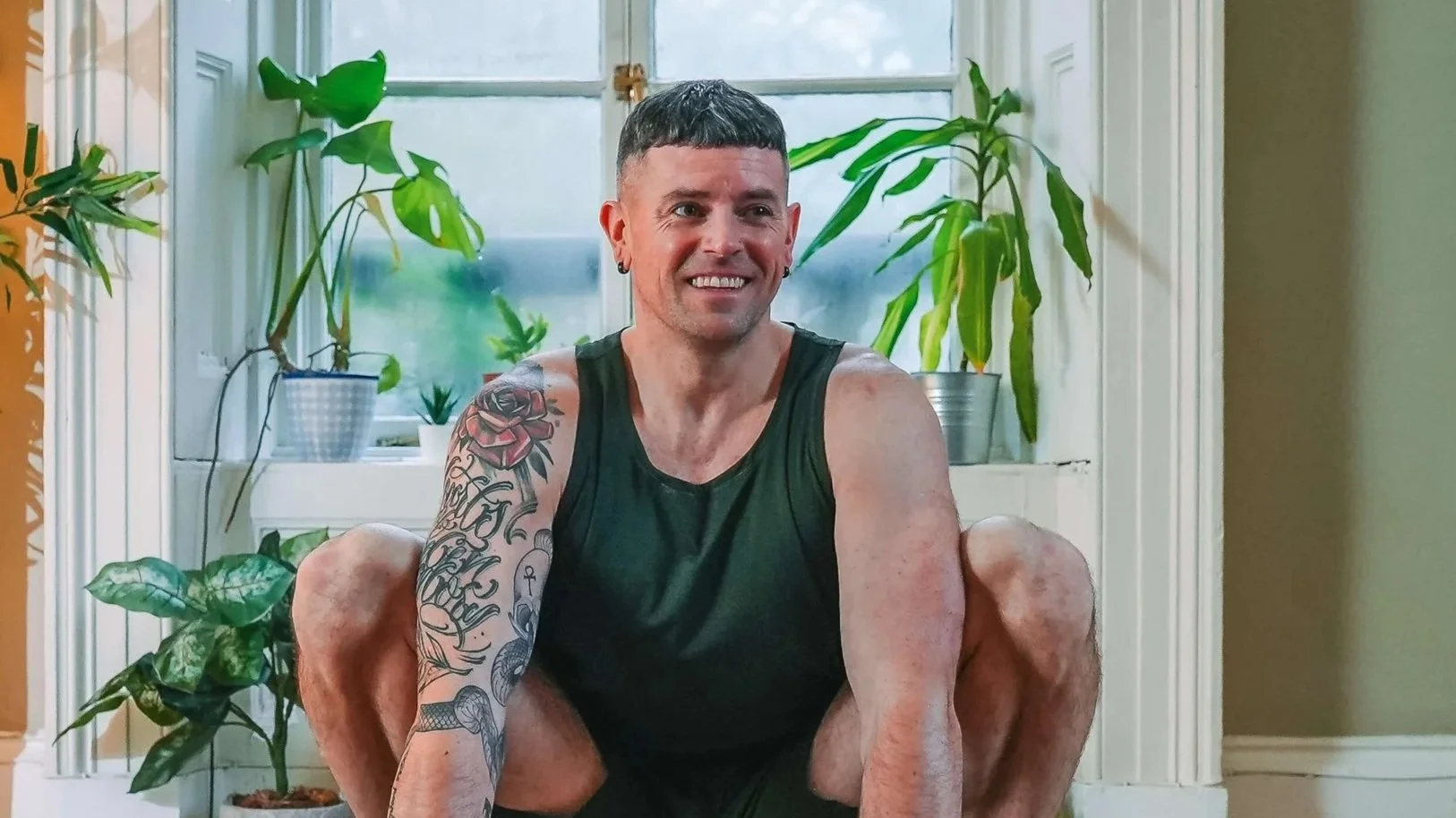 A young man with short dark hair, tattoos on his arm, and earrings, sitting with his knees up in a room filled with potted plants, smiling at the camera.