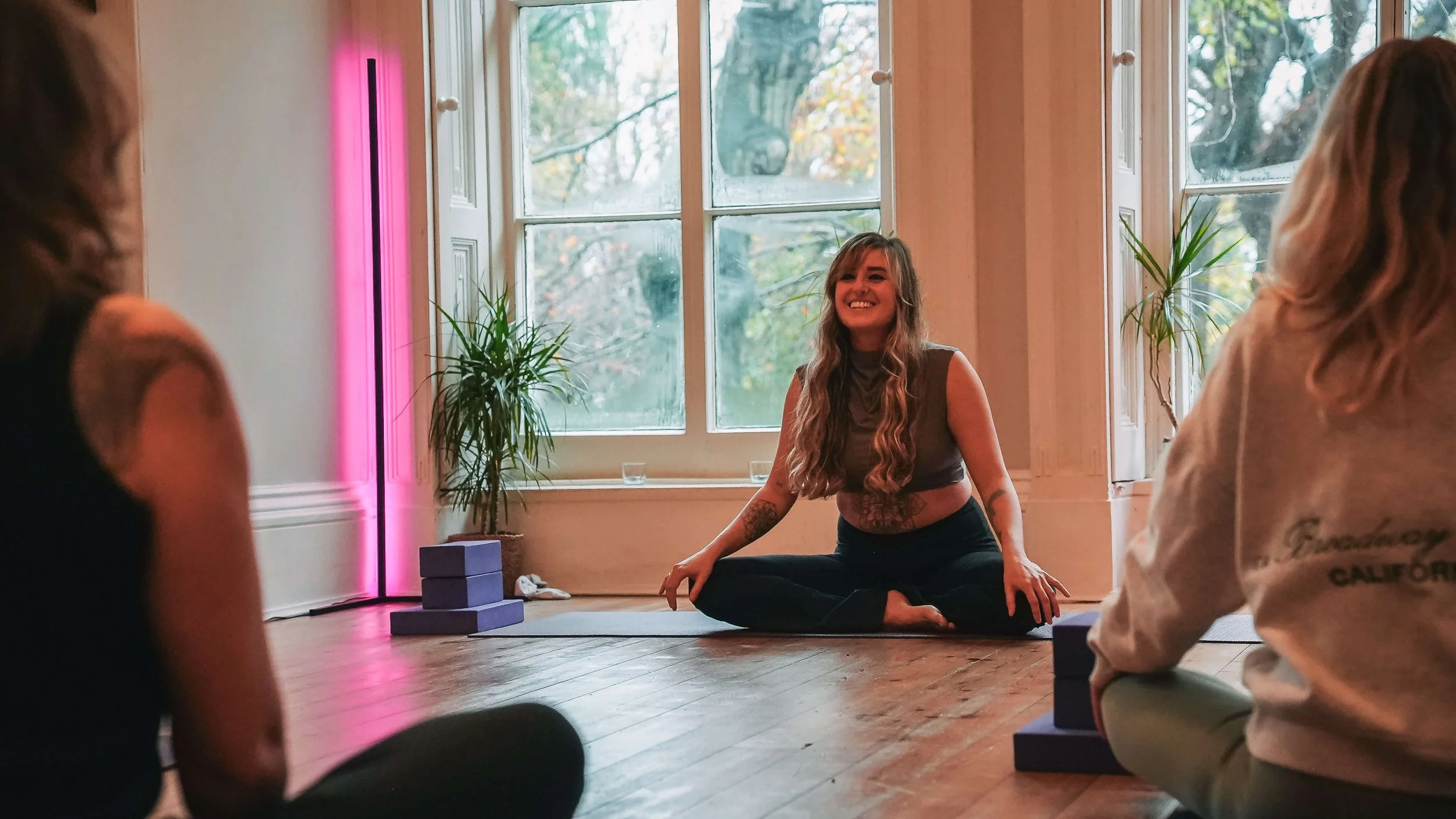 A woman with long wavy hair sitting cross-legged on the floor, smiling, leading a yoga or meditation session with a small group of people indoors near large windows with a view of trees.