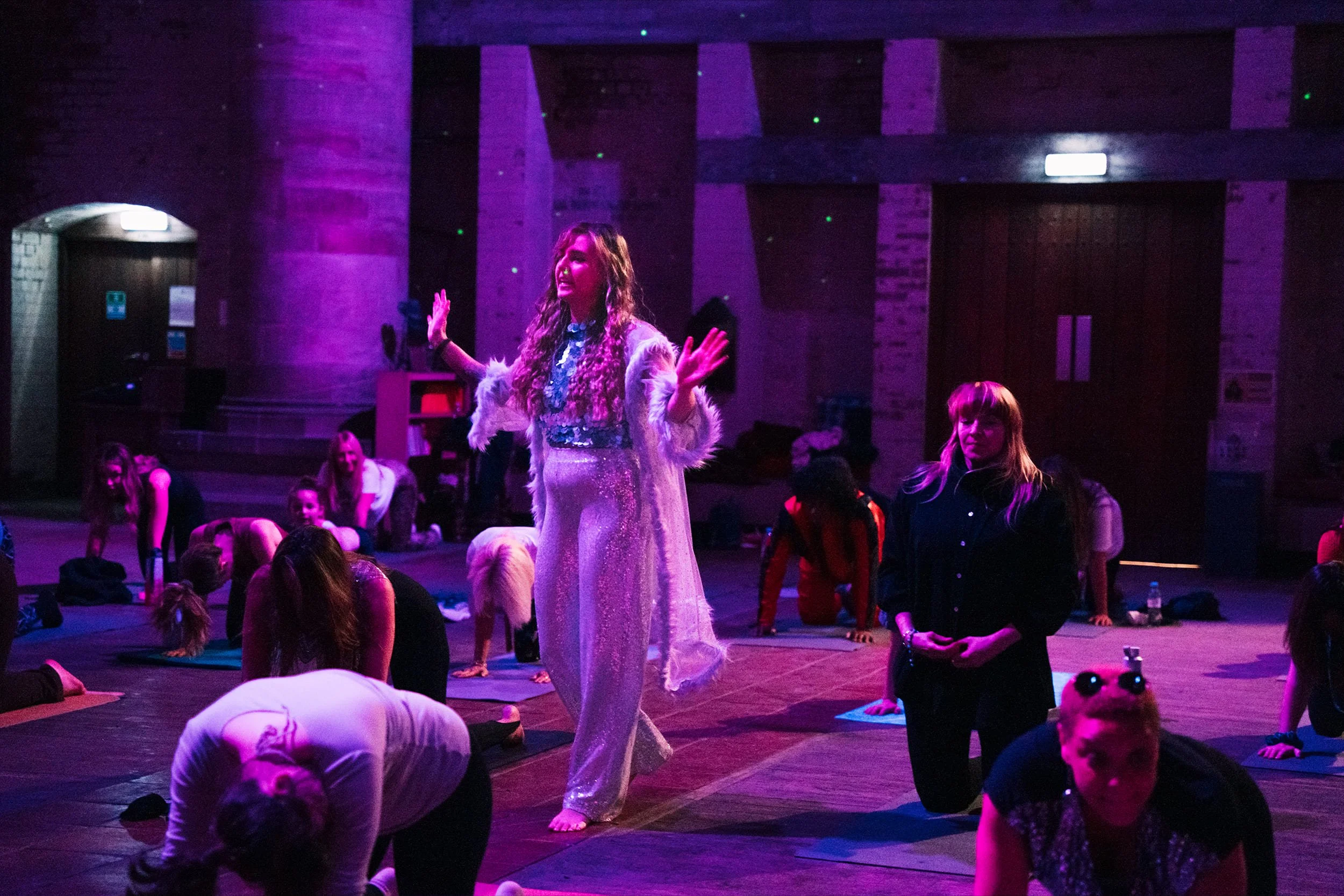 A woman in a sparkling dress and fluffy jacket leading a group in a yoga or dance class in a dimly lit room with purple lighting, with other people sitting or kneeling on the floor.